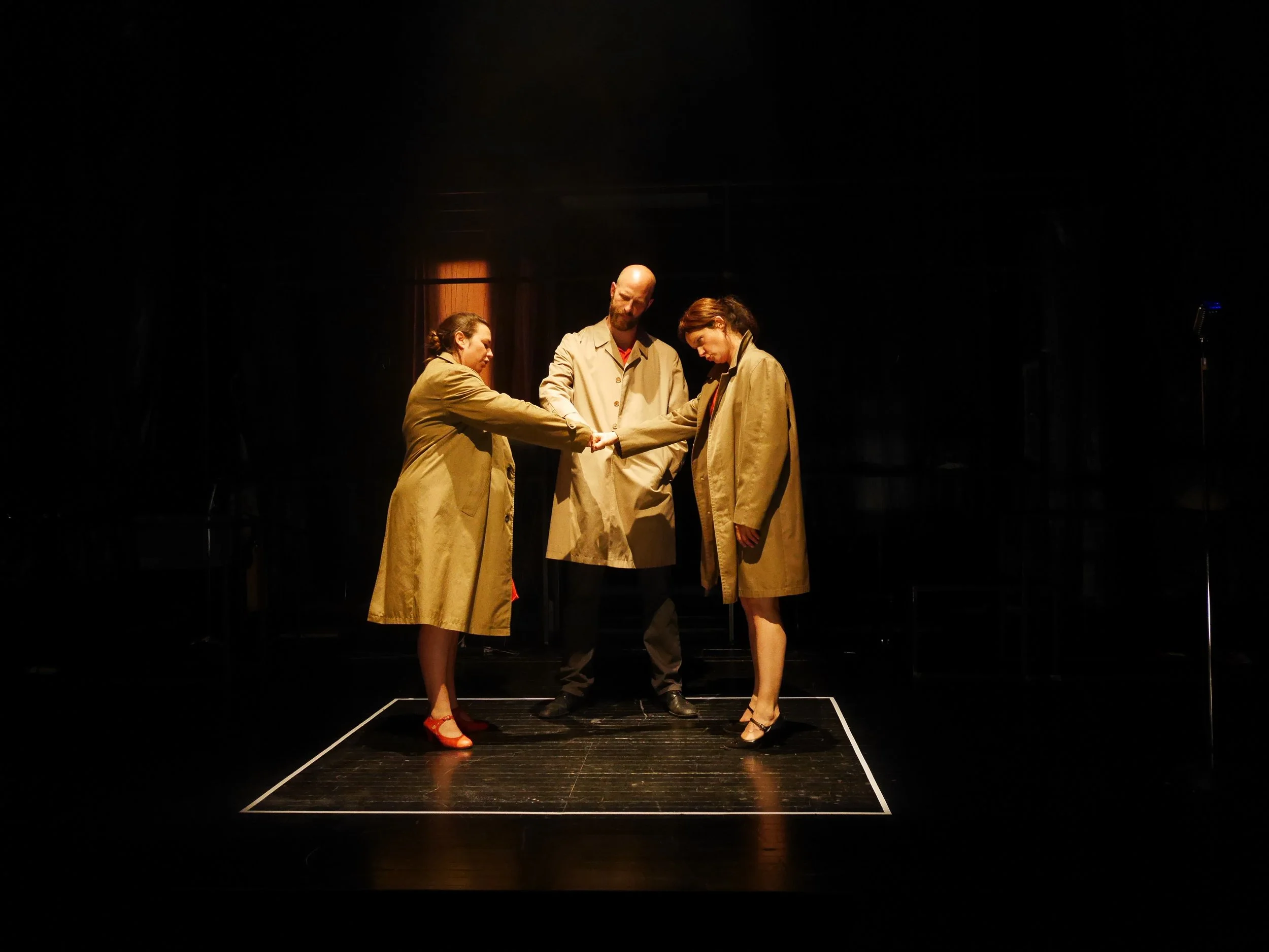 Three actors on stage, wearing beige trench coats, holding hands in a circle, under a spotlight with a dark background.