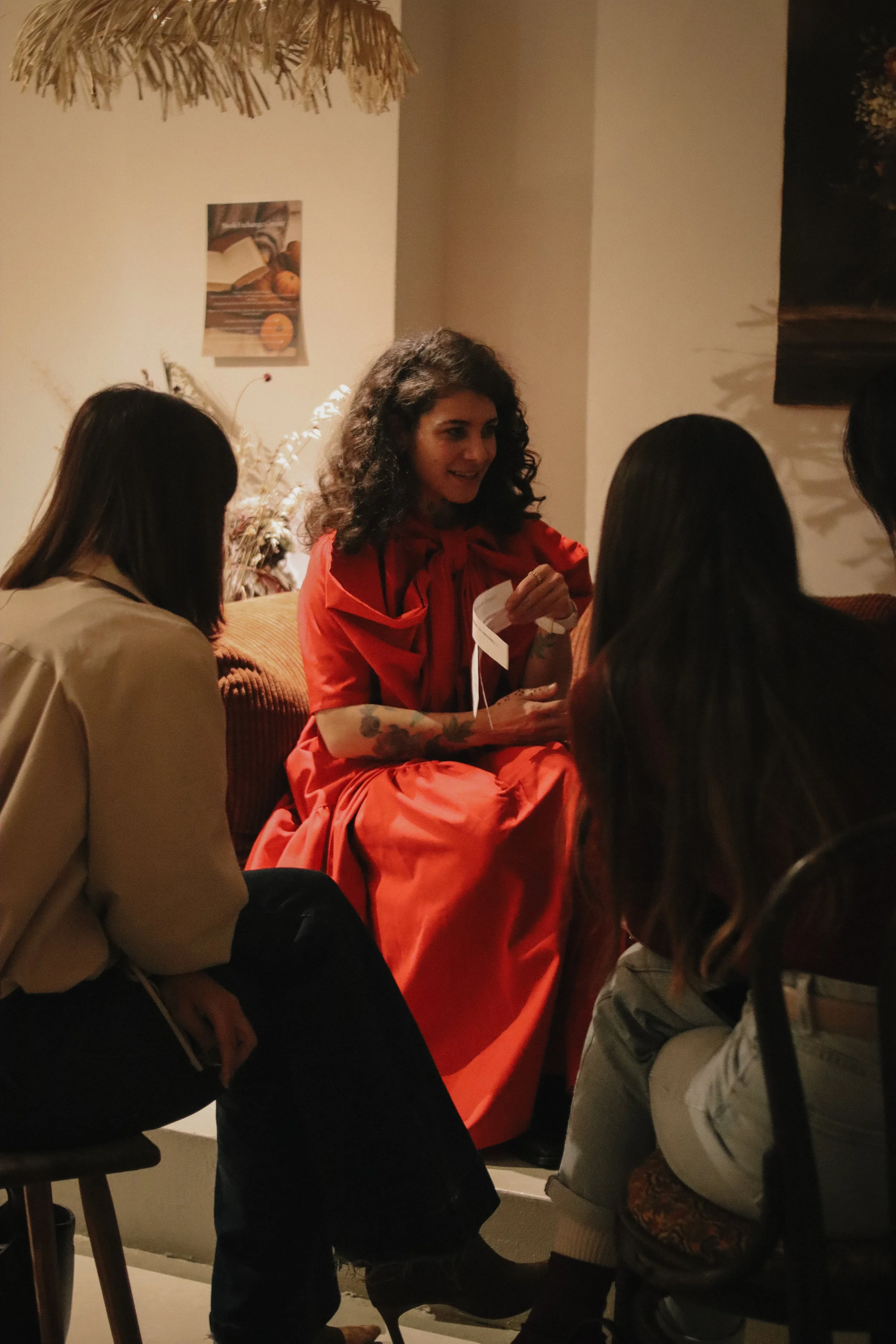 A woman in a red dress and curly hair is sitting on a sofa, engaging in conversation with three others. The setting appears cozy and warmly lit, possibly in a home or cafe.
