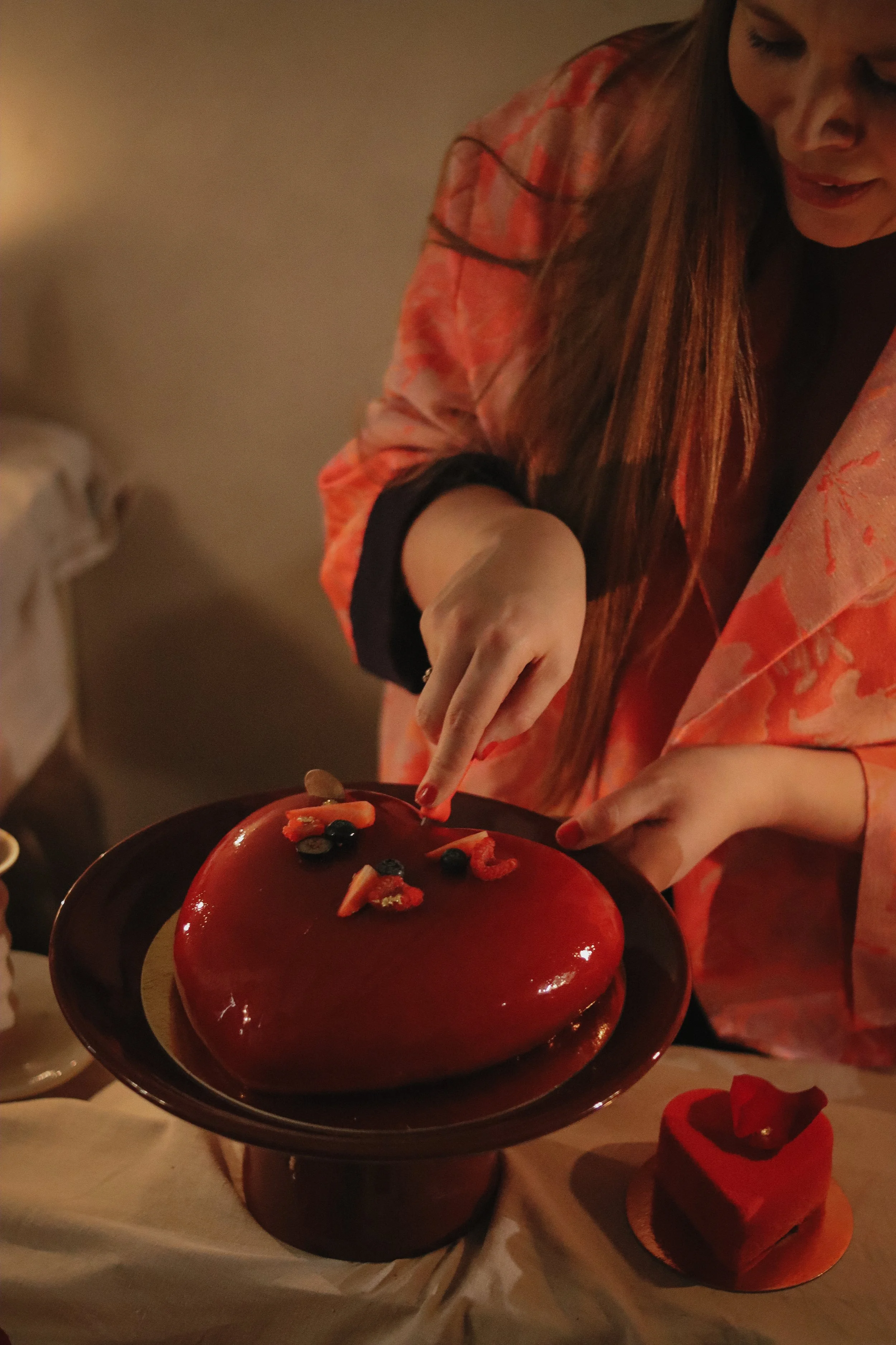 Woman with long red hair in a pink and orange floral robe decorates a heart-shaped cake with berries and strawberries.