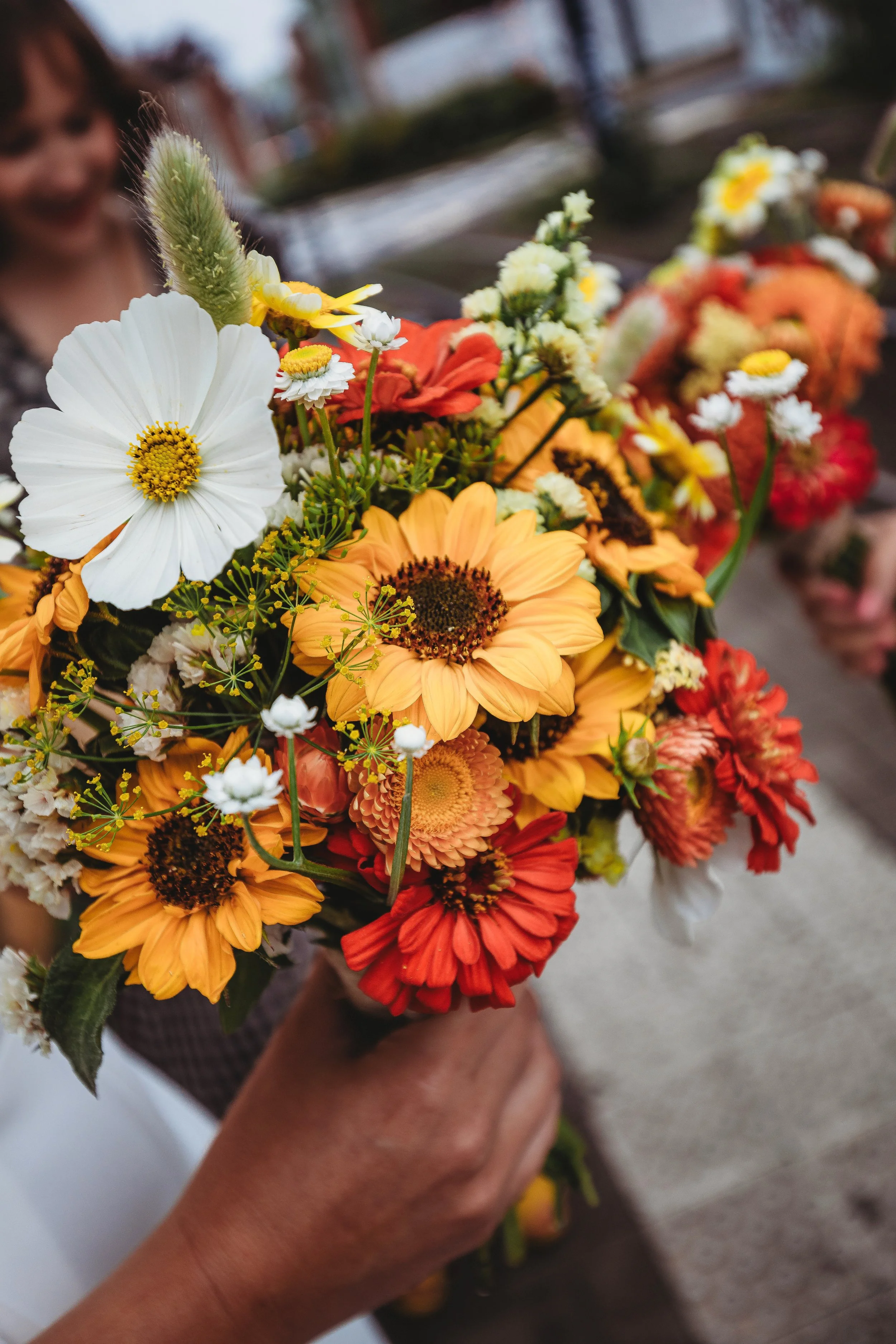 Person holding a colorful bouquet of mixed flowers, including sunflowers, daisies, and other vibrant blooms, with a blurred background.