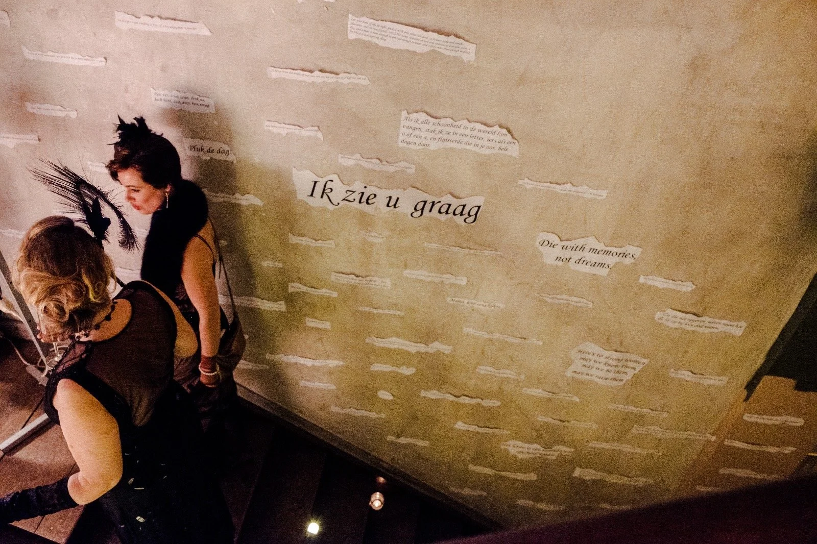 Two women with feathered headpieces looking at an exhibit or display on a wall with various texts in different languages and fonts.