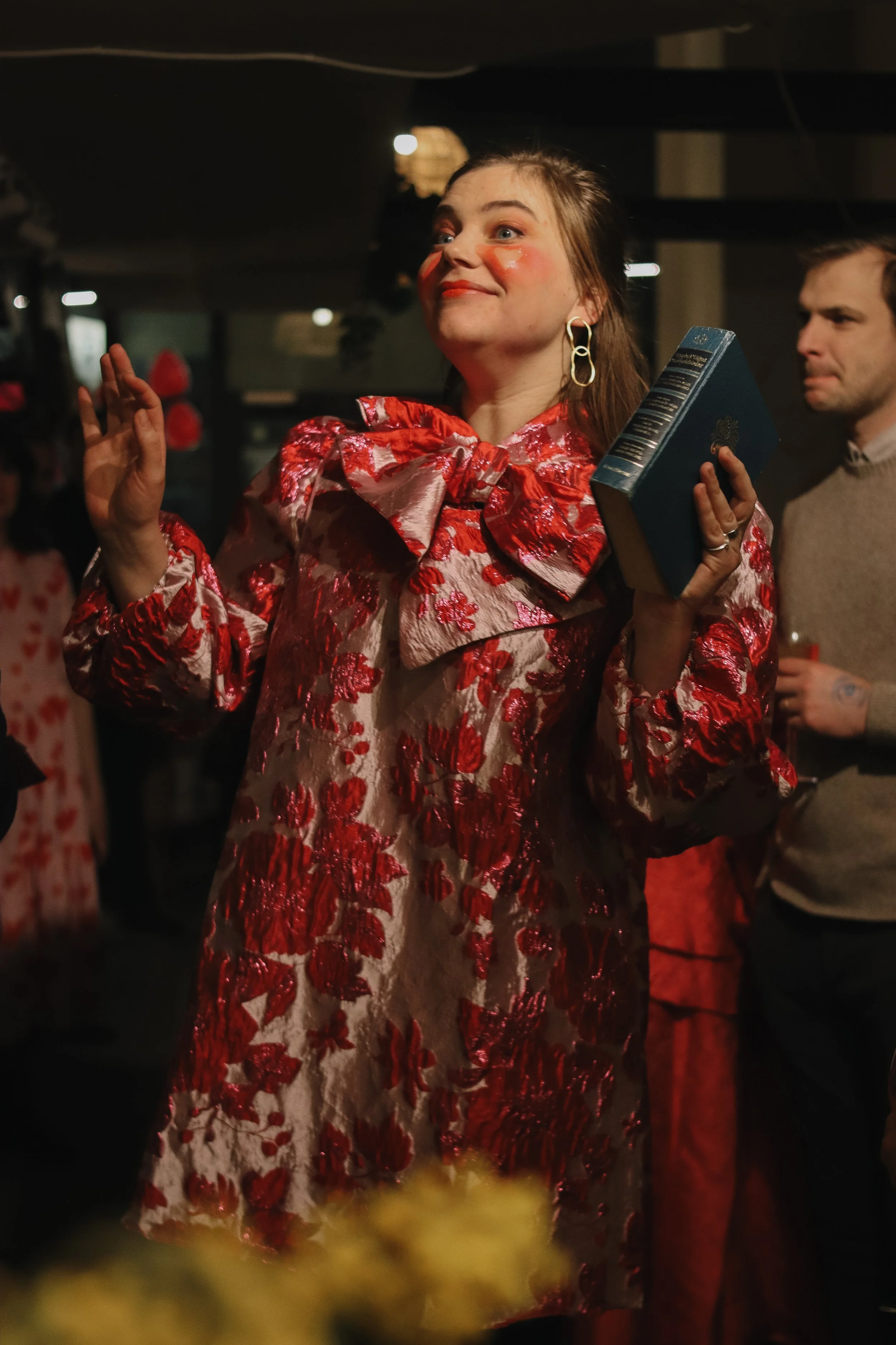 A woman dressed in a floral red and pink dress with a large bow around her neck, holding a blue book, standing in a crowded indoor setting.