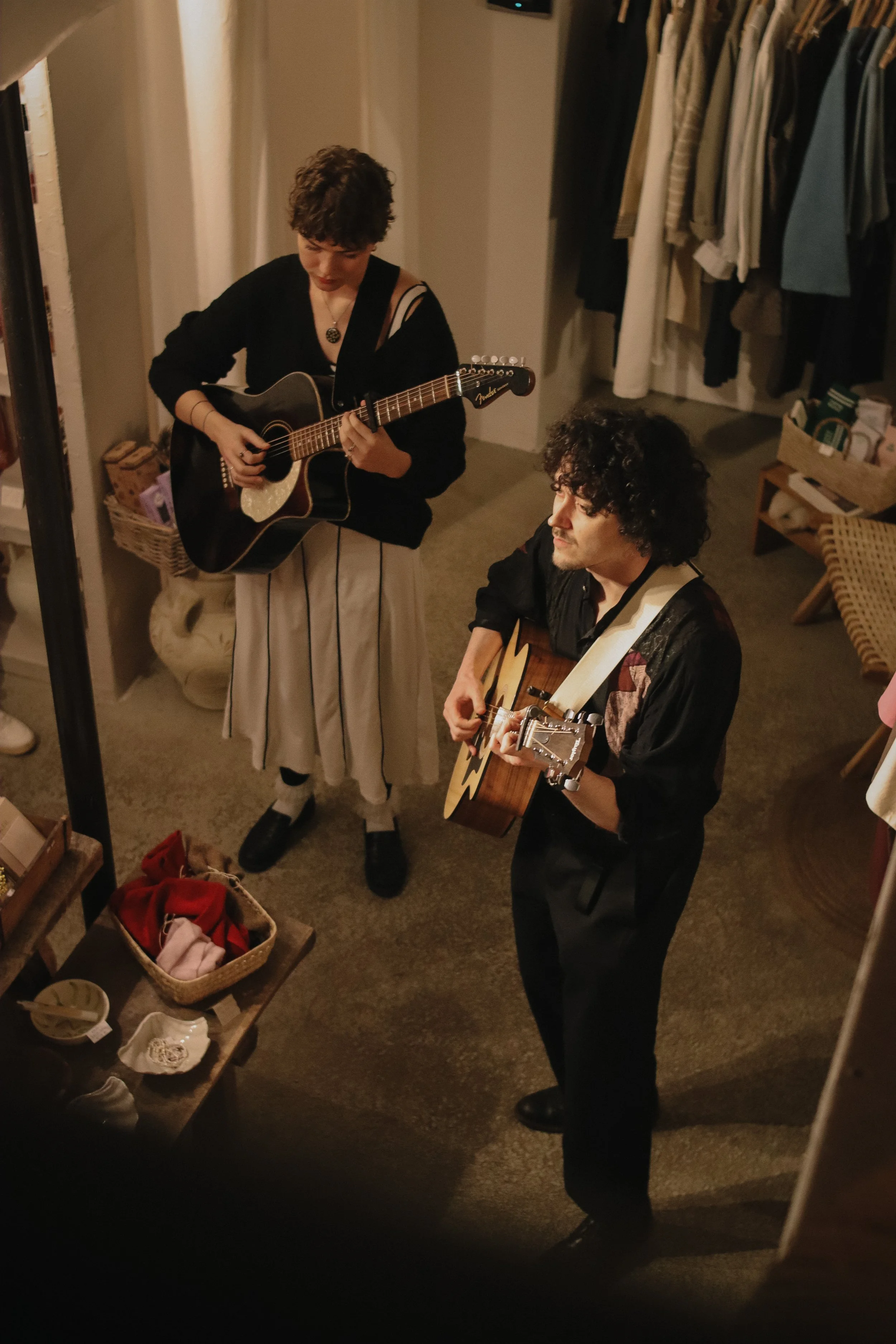 Two people playing acoustic guitars in a cozy indoor setting, surrounded by clothing and household items.