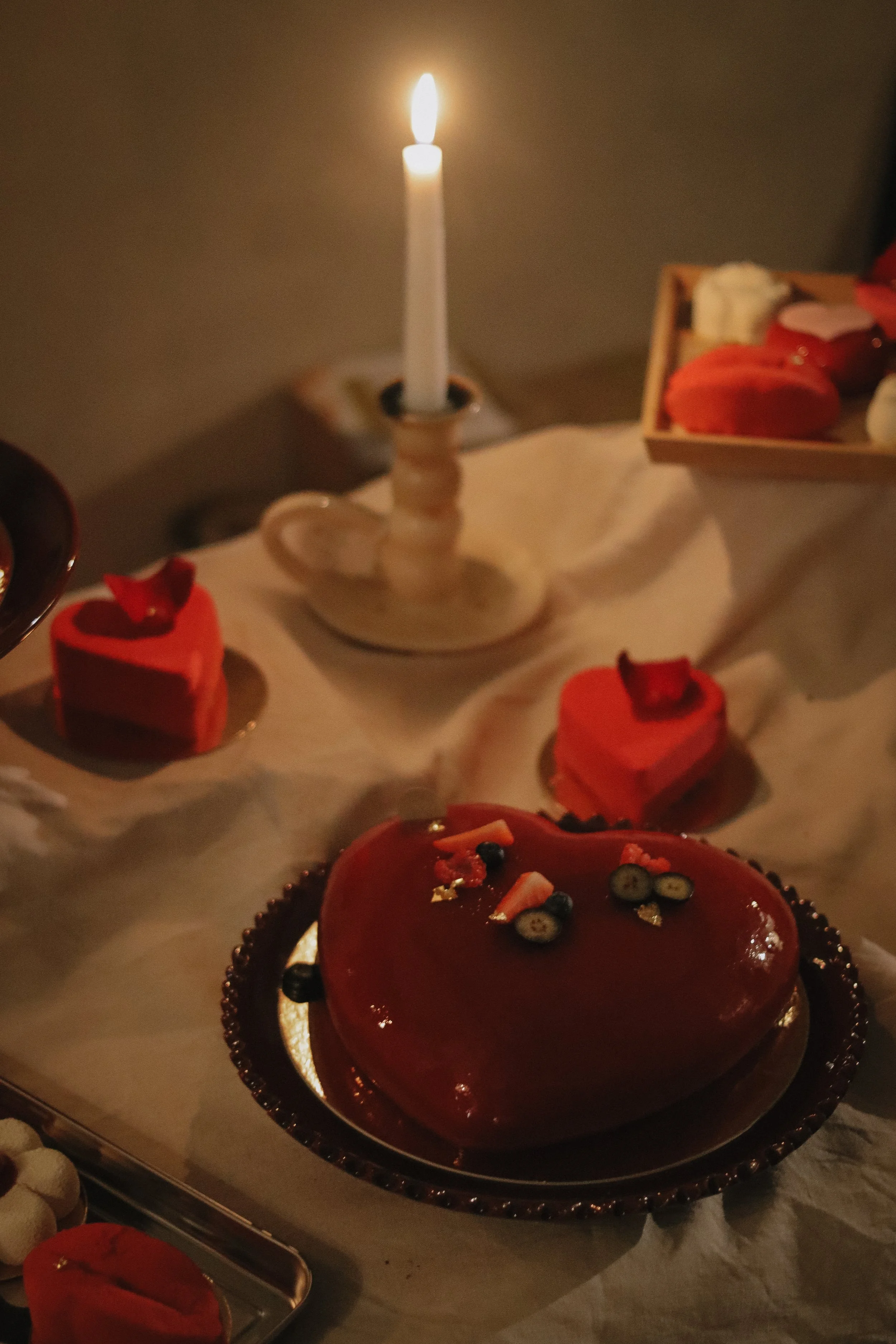 A heart-shaped red cake with fruit toppings, surrounded by small red and white desserts, illuminated by a lit candle on a table.