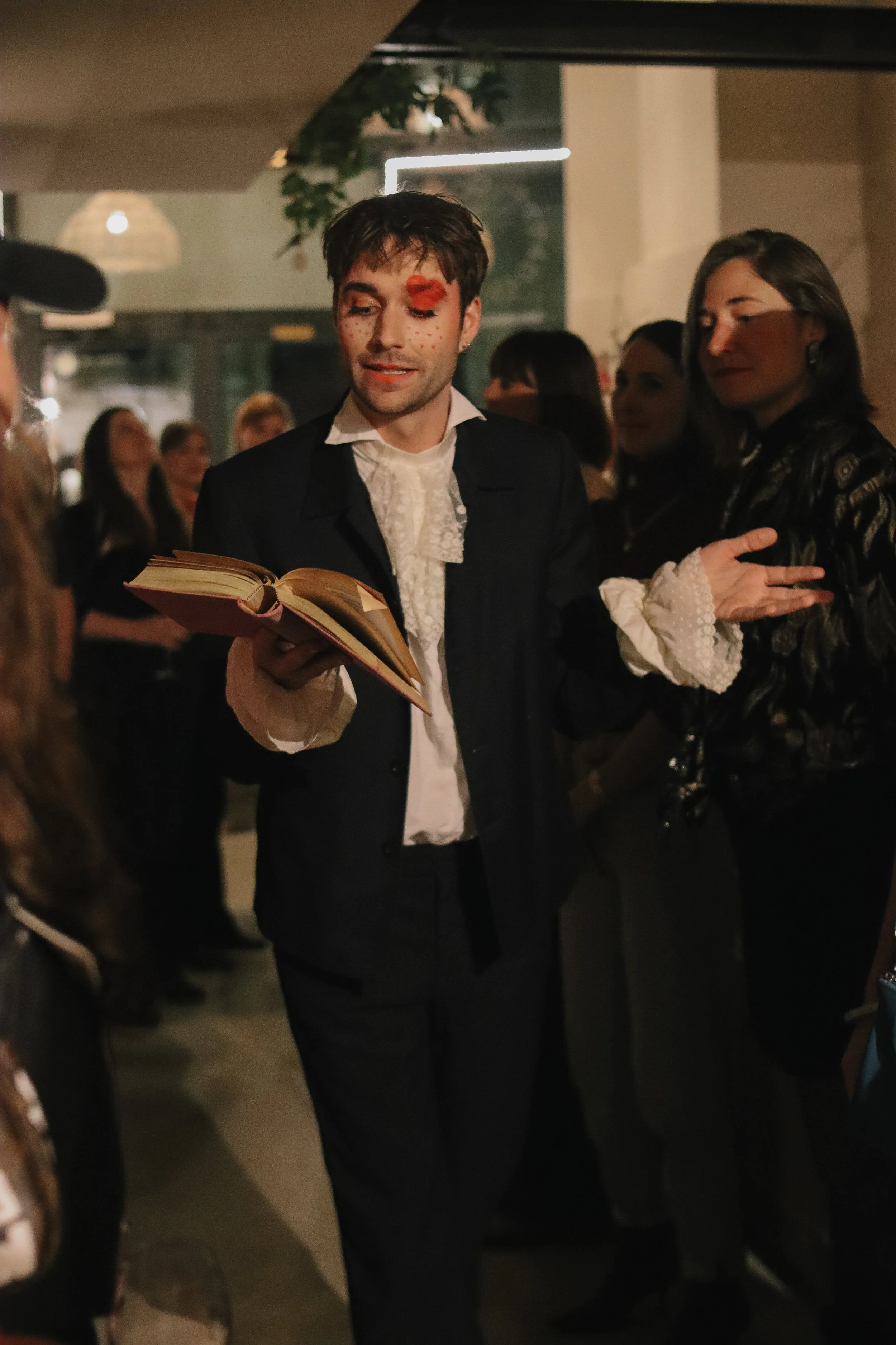 A young man dressed in historical clothing, holding an open book, standing in a dimly lit social setting with other people in the background.