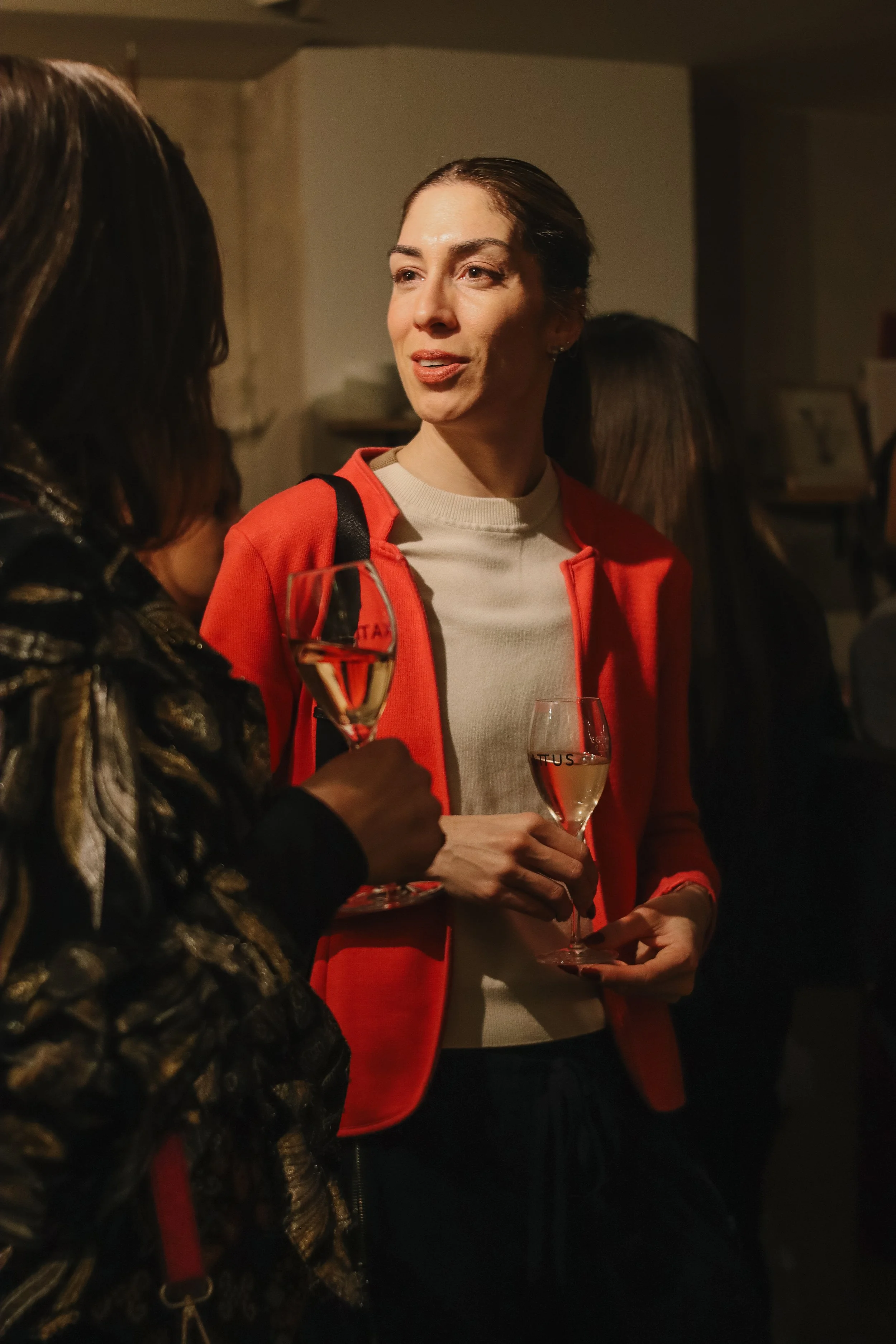 Two women having a conversation at a social gathering, holding glasses of champagne, in a warmly lit indoor setting.