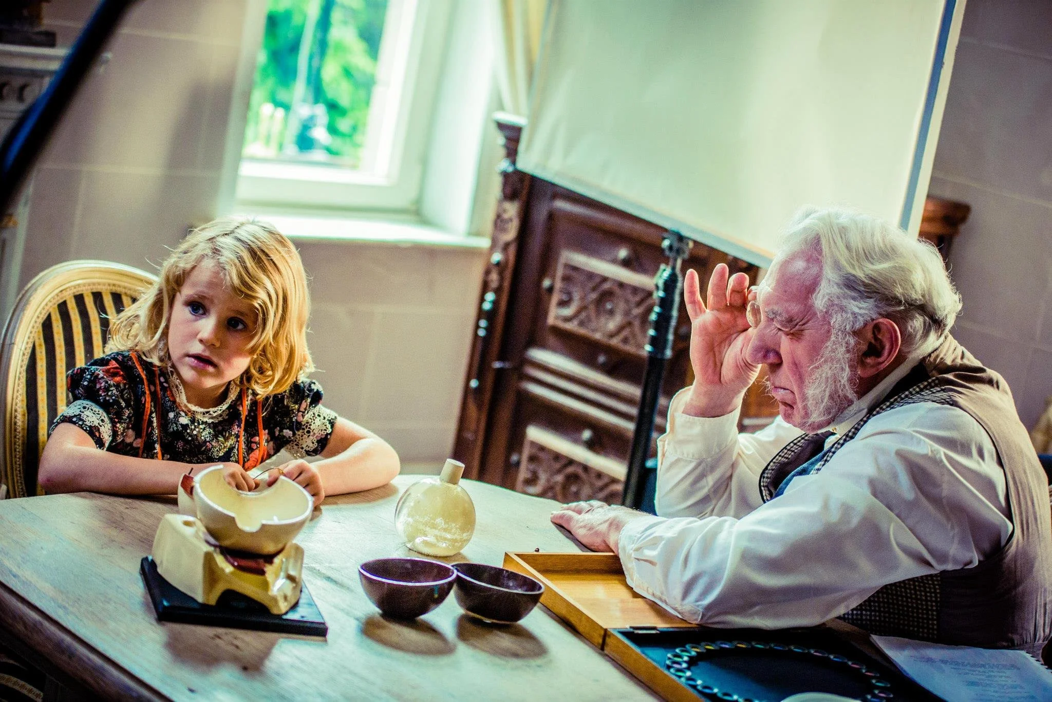 A young girl with blonde hair sitting at a wooden table with a puzzled or concerned expression on her face, next to an elderly man with white hair and sideburns, who appears to be rubbing his eyes or face in distress. The setting is a room with a win