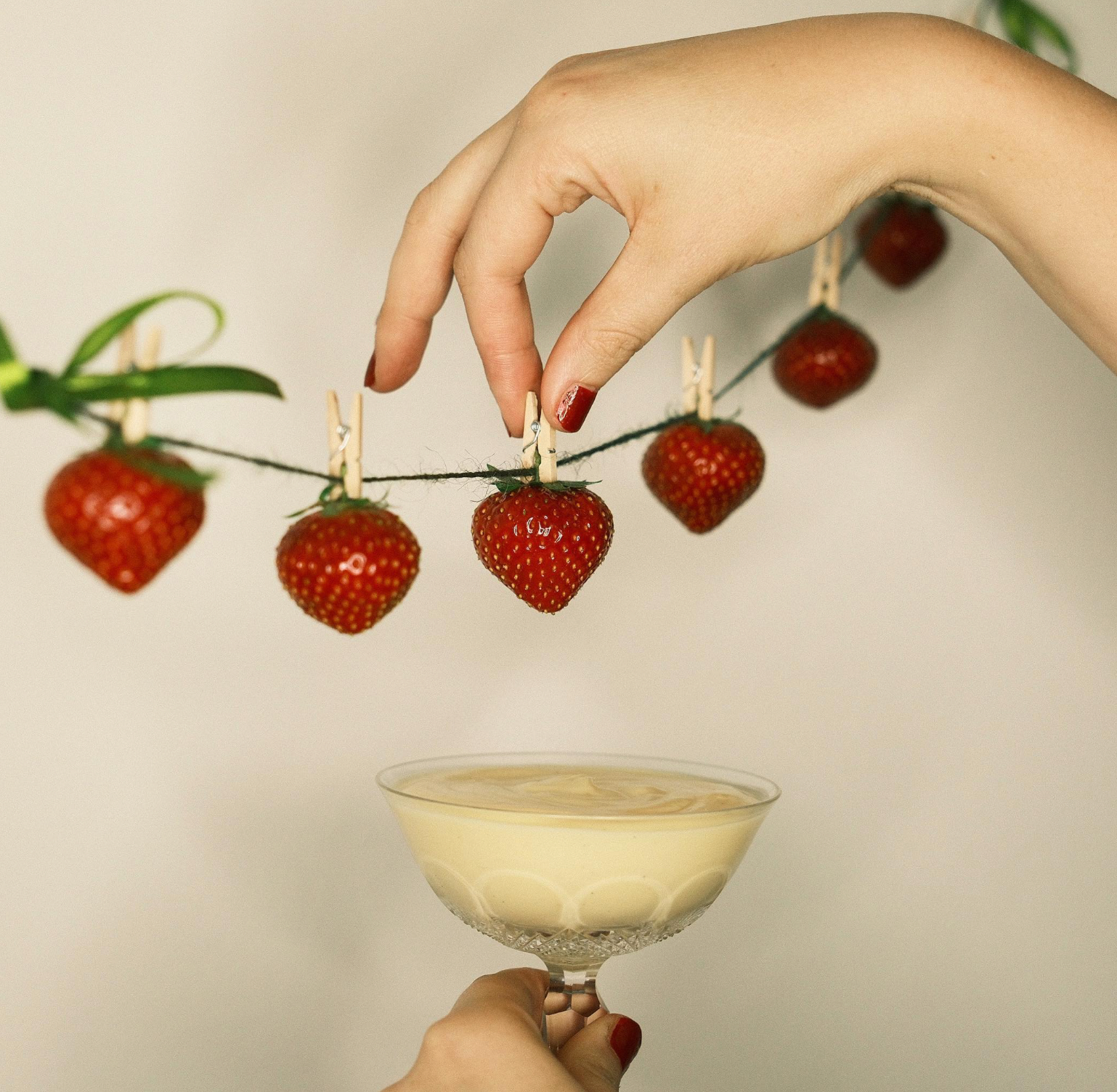 A hand with red-painted nails holding a glass of creamy drink beneath a string of strawberries clipped to a line, with another hand holding the glass.