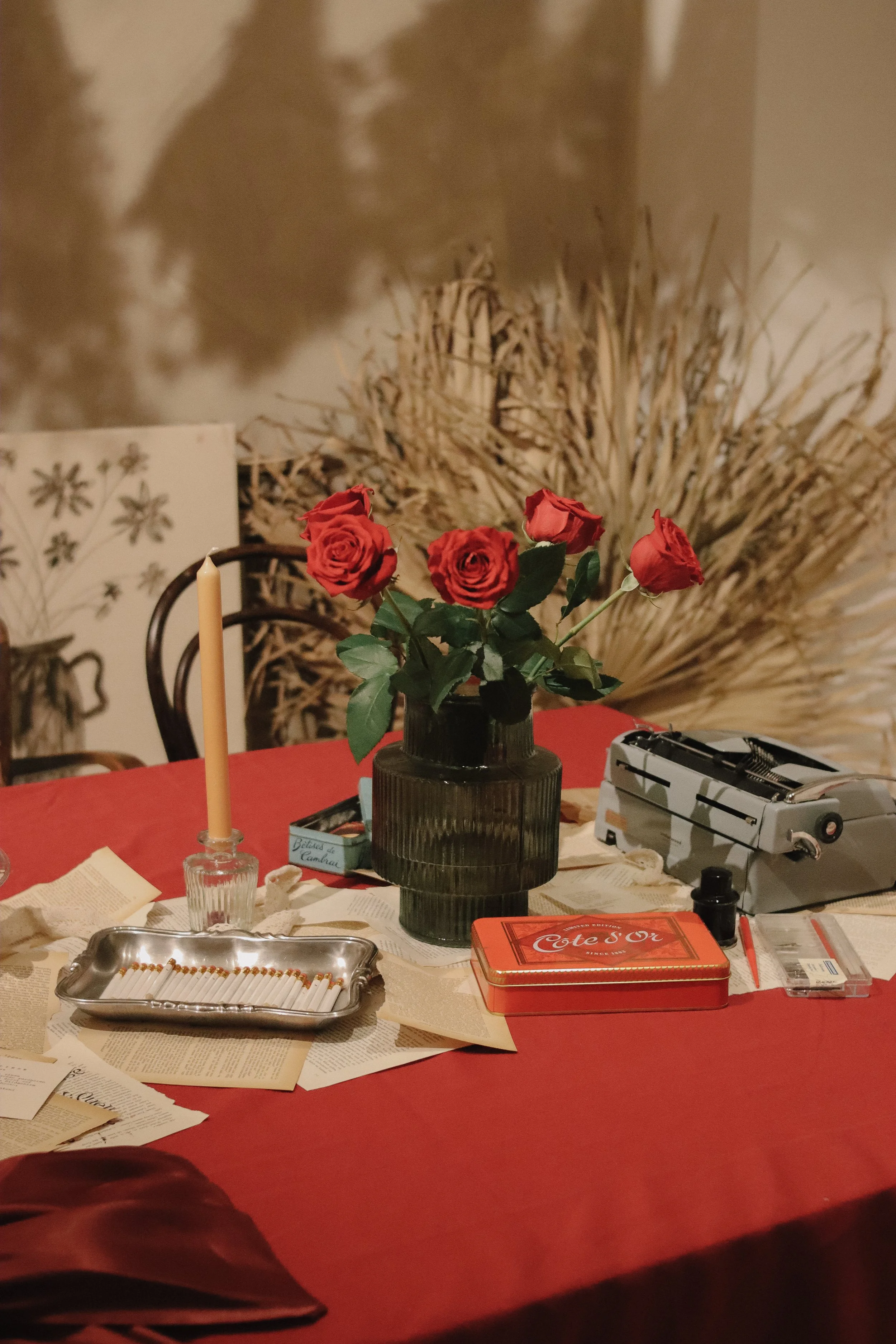 A table with red tablecloth, vase with red roses, scattered papers, an orange cigarette tin, and an old typewriter, with a candle in a glass holder and a decorative backdrop.