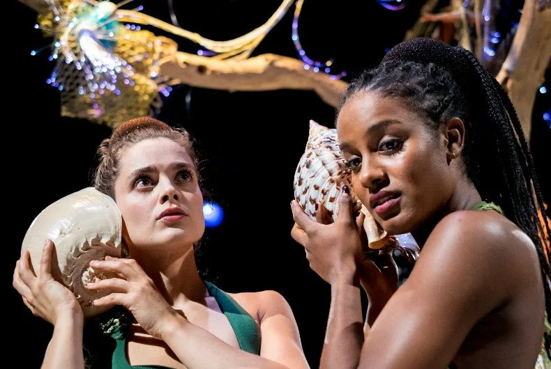 Two women holding seashells on their shoulders, looking contemplative, with decorative lights in the background.