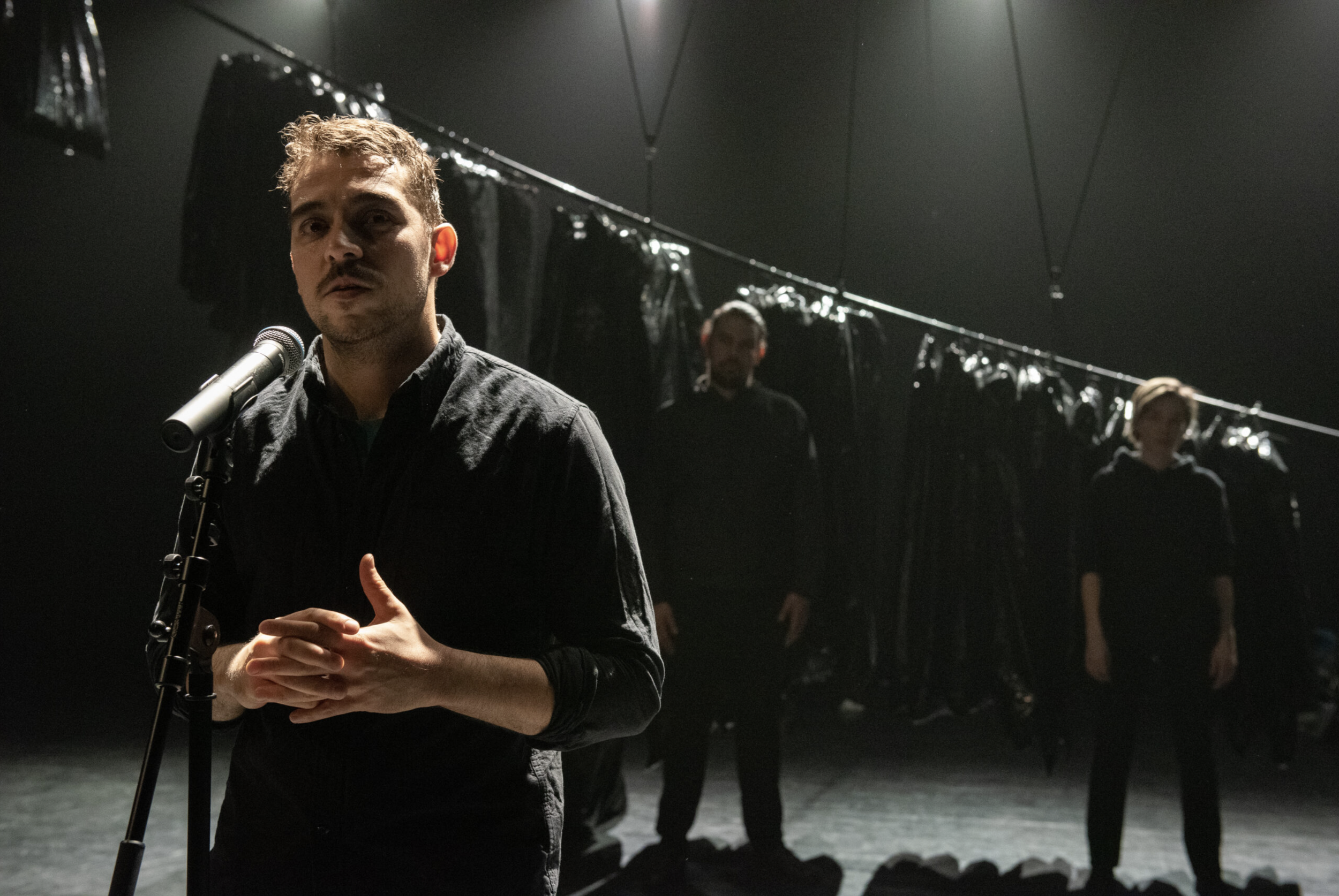 A man speaking into a microphone on stage with two people and a rack of clothing or costumes in the background.