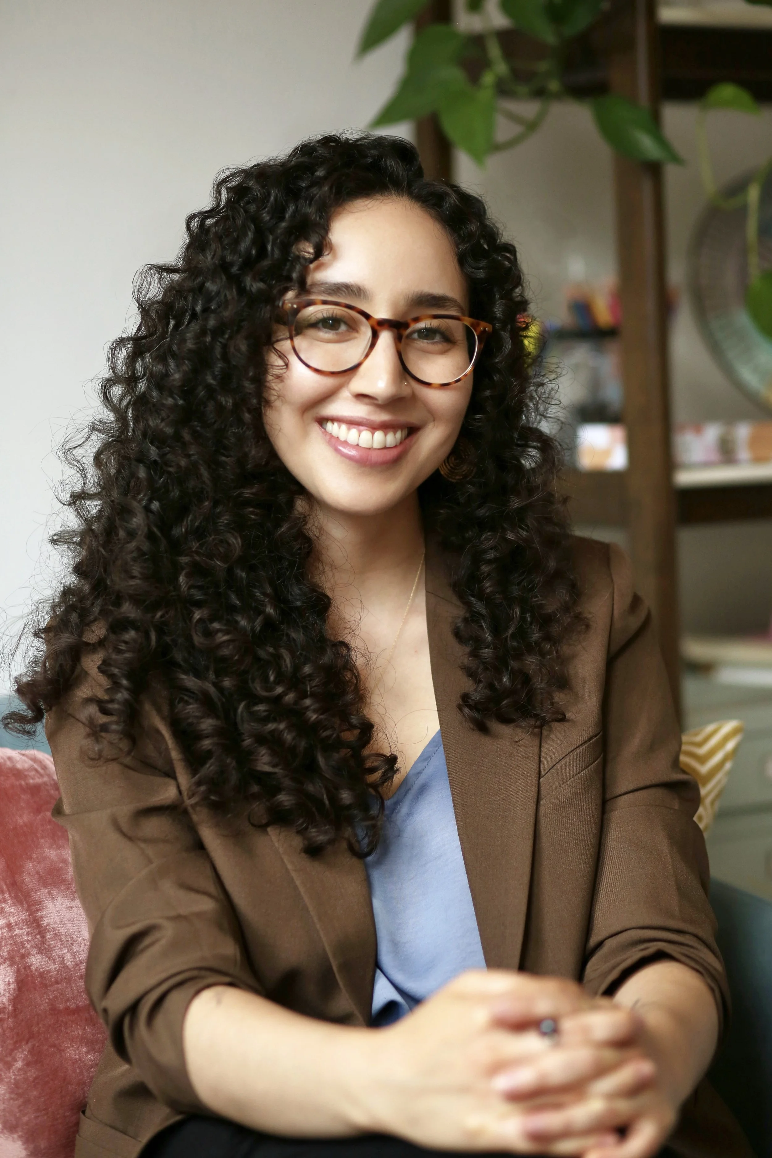 A woman with curly dark hair, glasses, and a nose piercing smiling with arms crossed, standing outside in front of a brick wall.