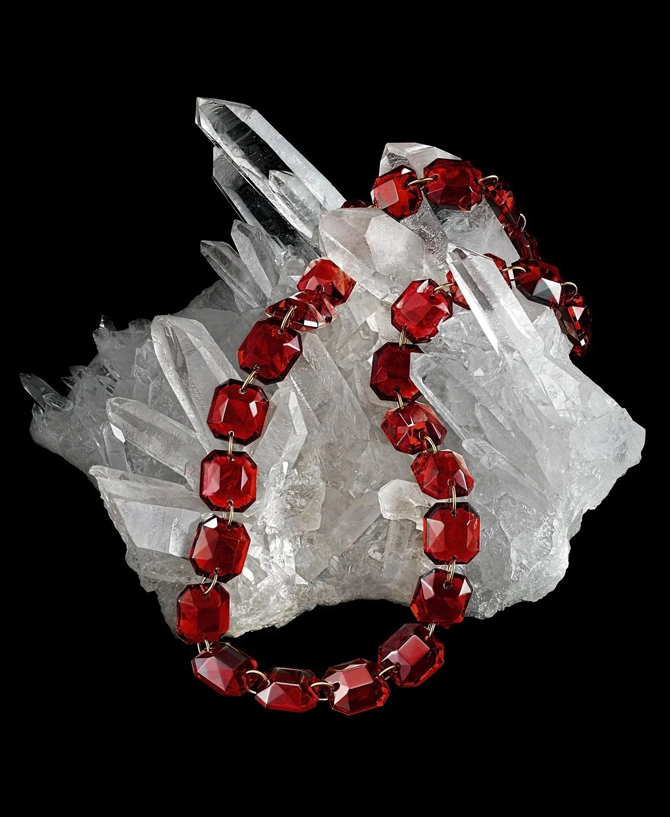 A clear red bead necklace draped over a cluster of clear quartz crystals on a black background.