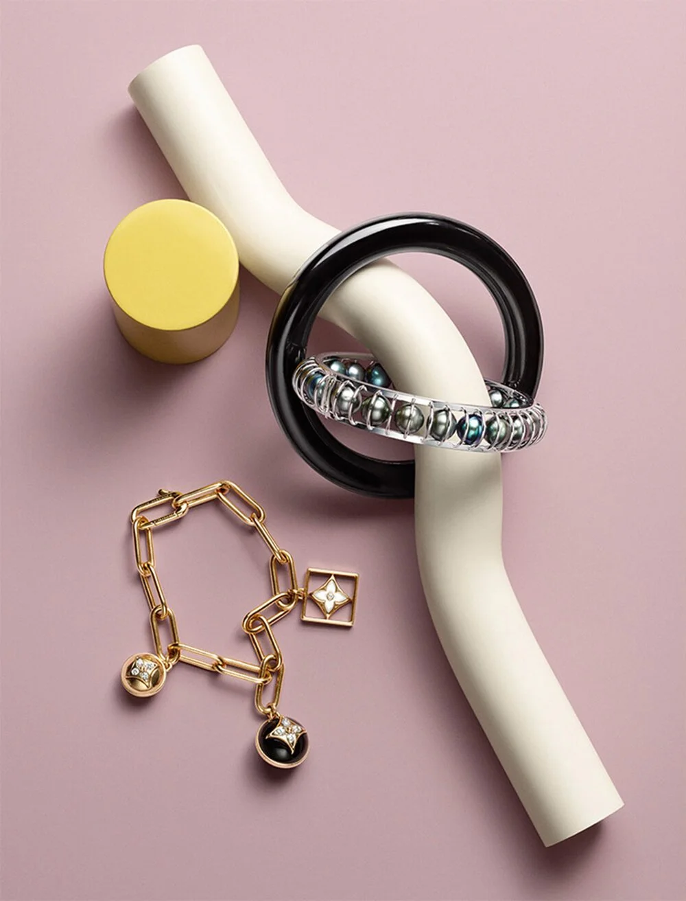 Various jewelry pieces placed on a pink background, including a yellow cylindrical object, a white curved structure, a black circular ring, a silver beaded bracelet, and a gold chain necklace with pendants.