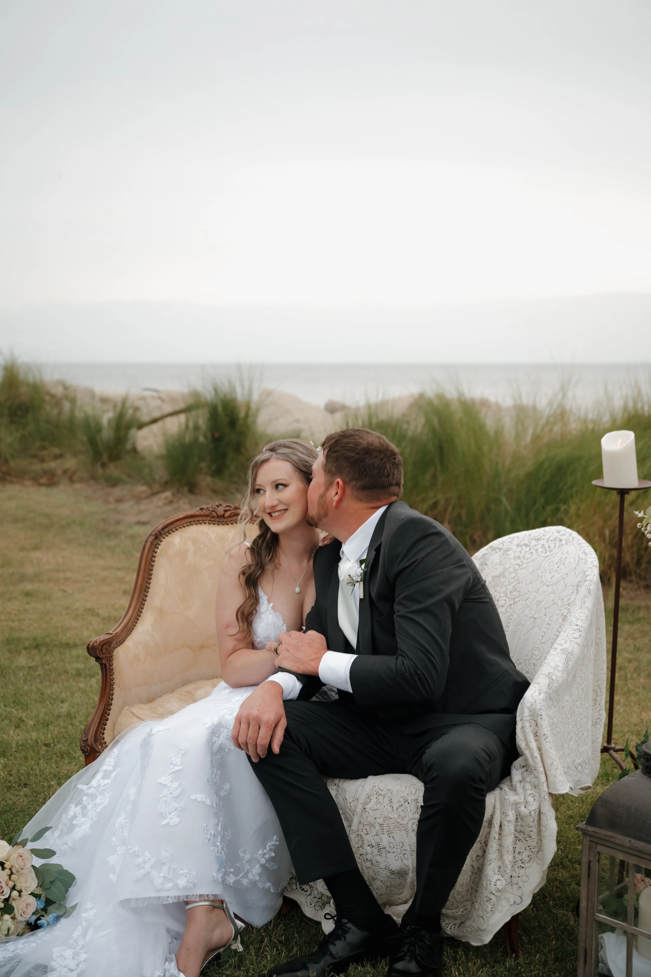 A bride and groom sitting on a vintage sofa outdoors at their wedding on a grassy area near the beach, with dunes and the ocean in the background. The groom is kissing the bride on the cheek, and she is smiling.