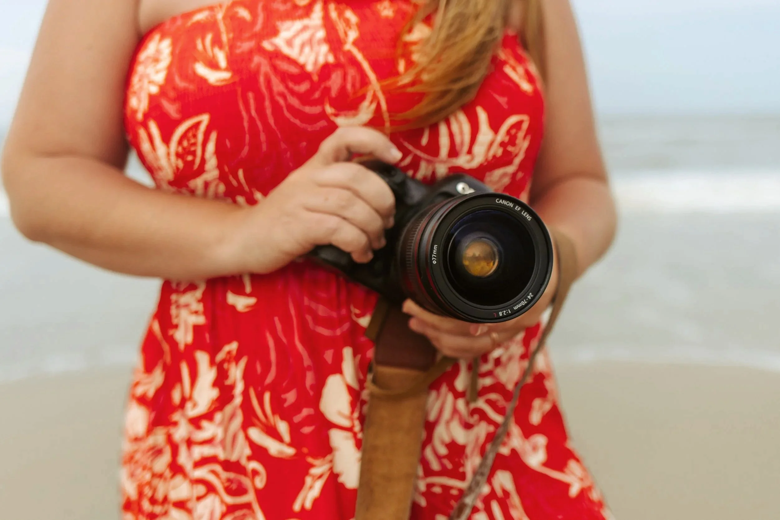 Person wearing a red dress with a floral pattern holding a black Canon camera with a large lens.