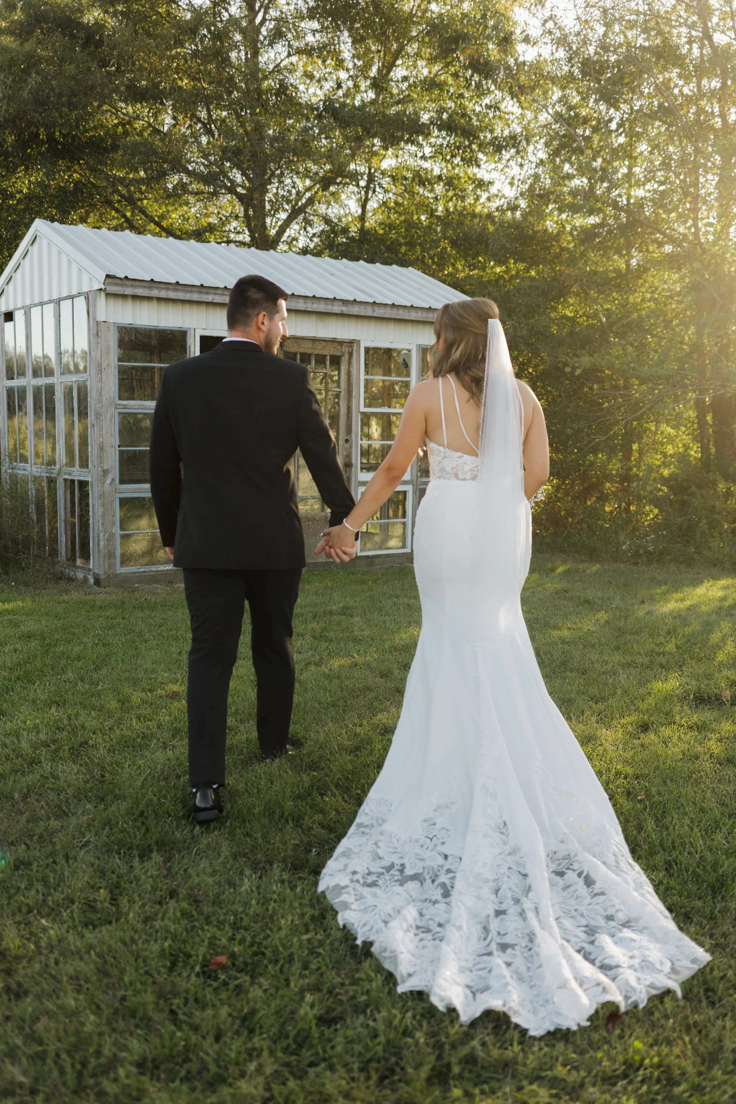 A bride and groom holding hands walking away from a small greenhouse on a grassy lawn during sunset.