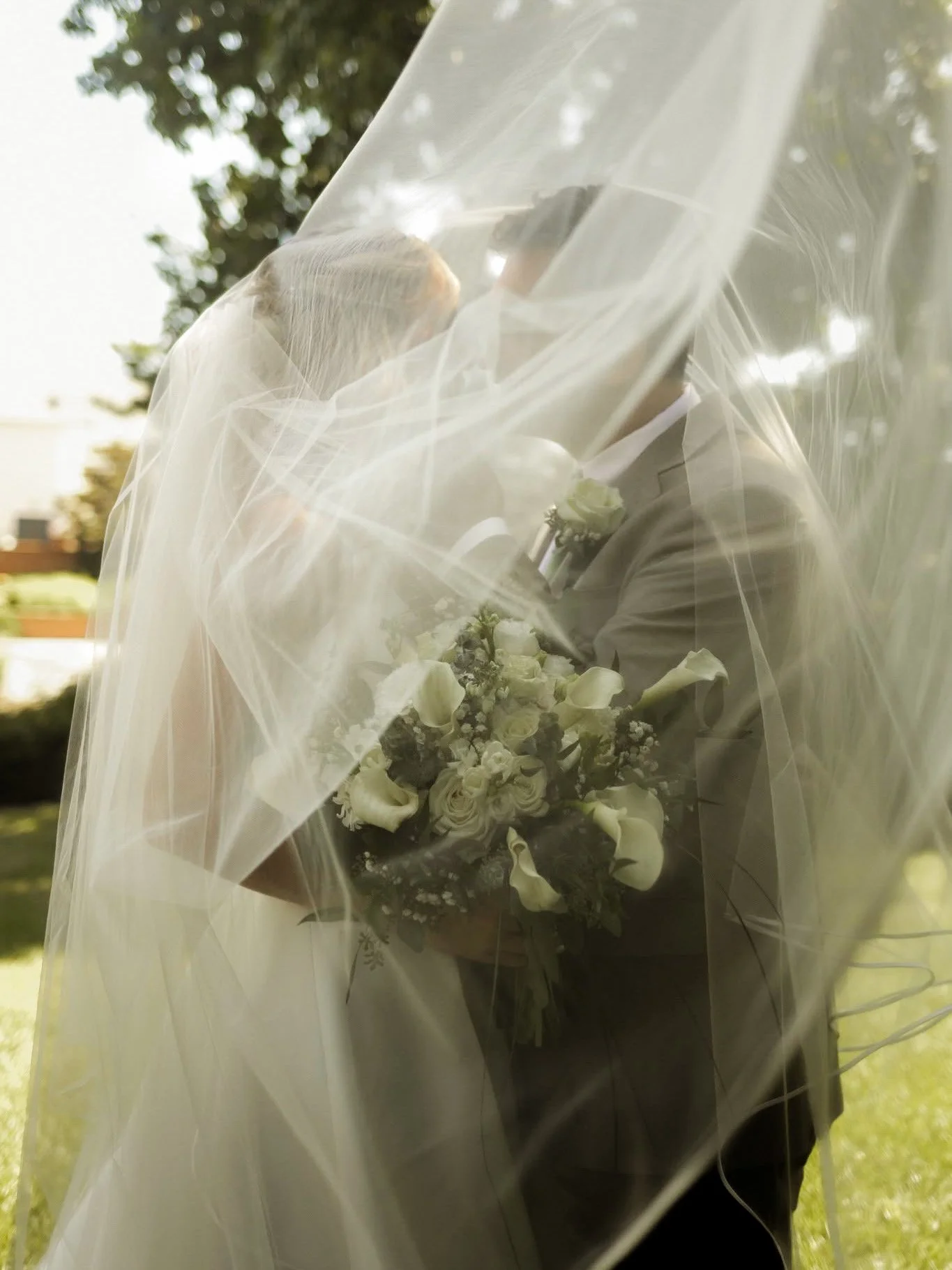 A wedding couple, partly obscured by a sheer veil, facing each other outdoors, with the bride holding a bouquet of white flowers.