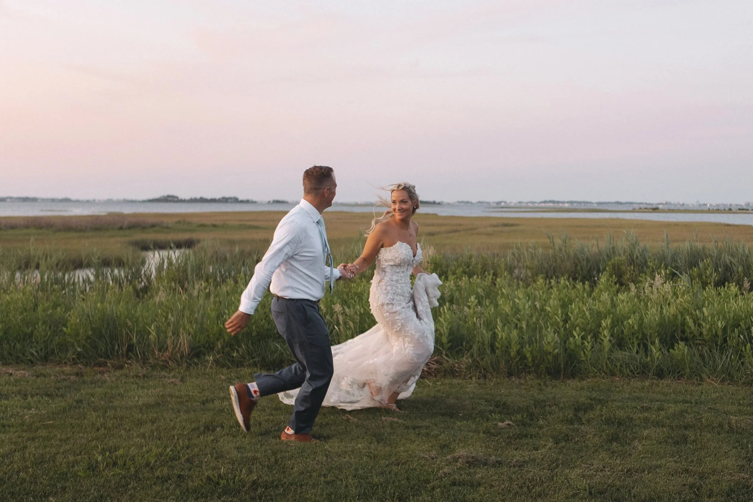 A bride and groom holding hands and smiling, running on grass near a marshland at sunset.
