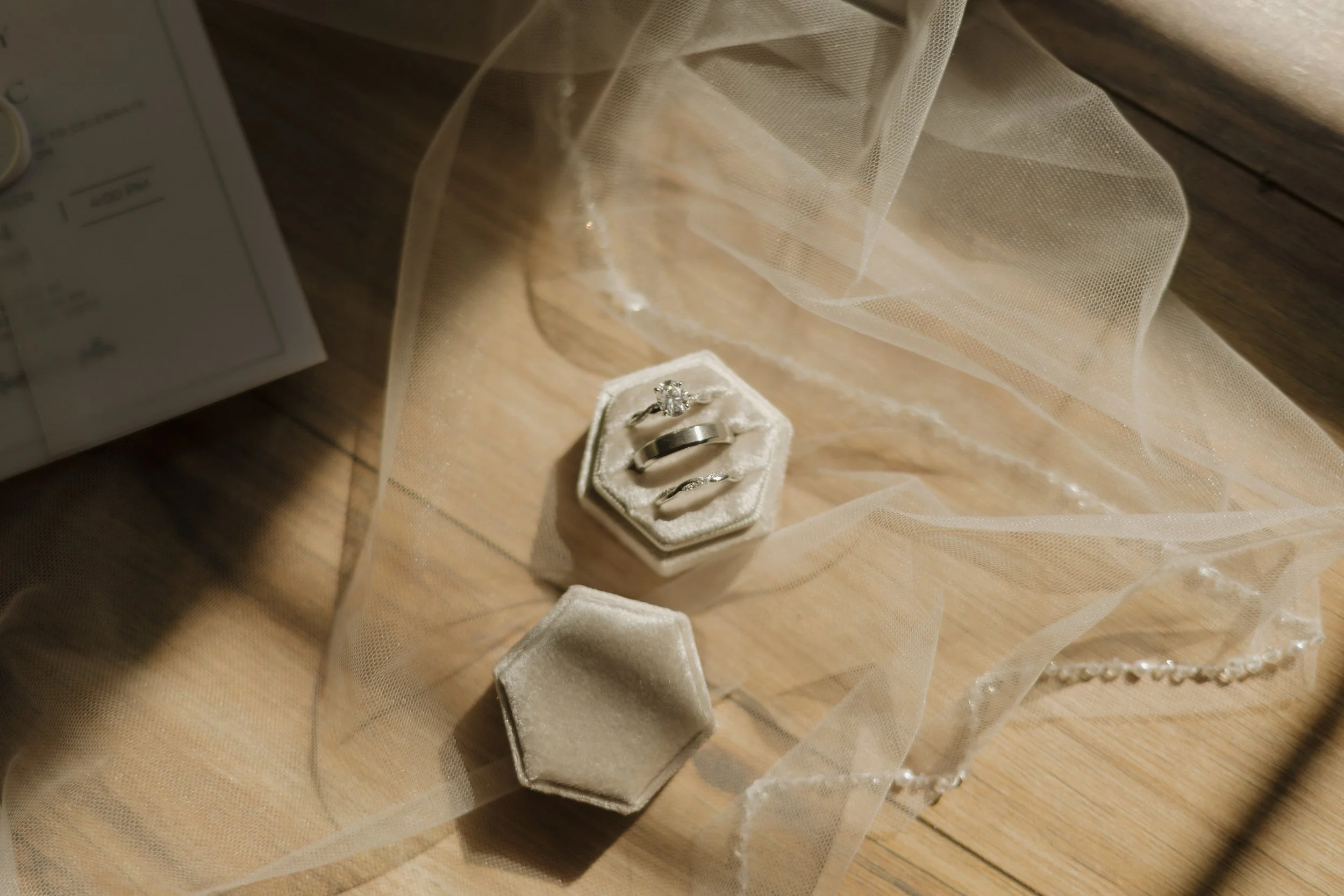Engagement or wedding rings inside a velvet ring box, with a pearl necklace and decorative tulle fabric on a wooden surface.