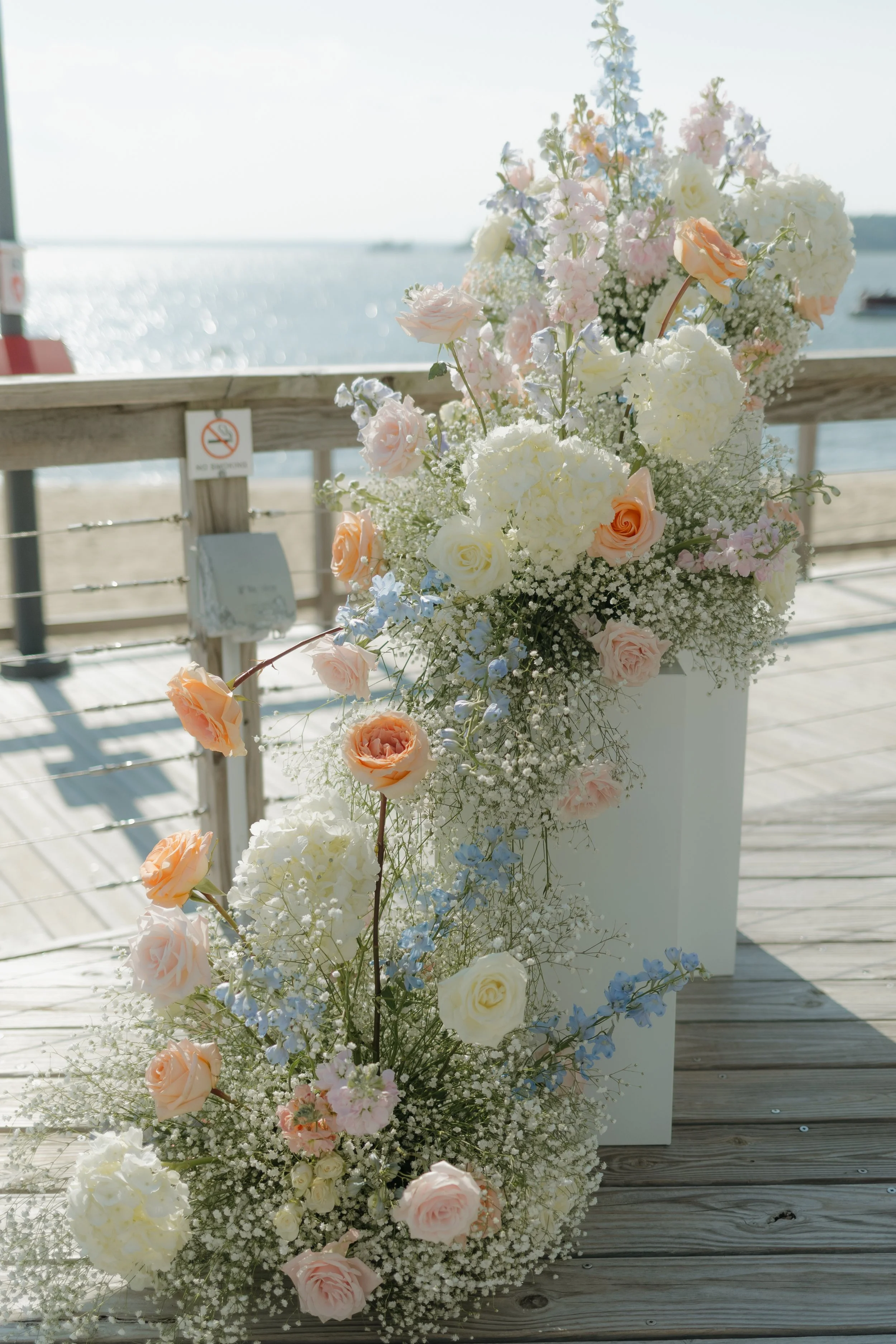 A large floral arrangement of pink, white, and blue flowers on a wooden deck near water. Coastal wedding, wedding details, ceremony florals, travel destination wedding photographer