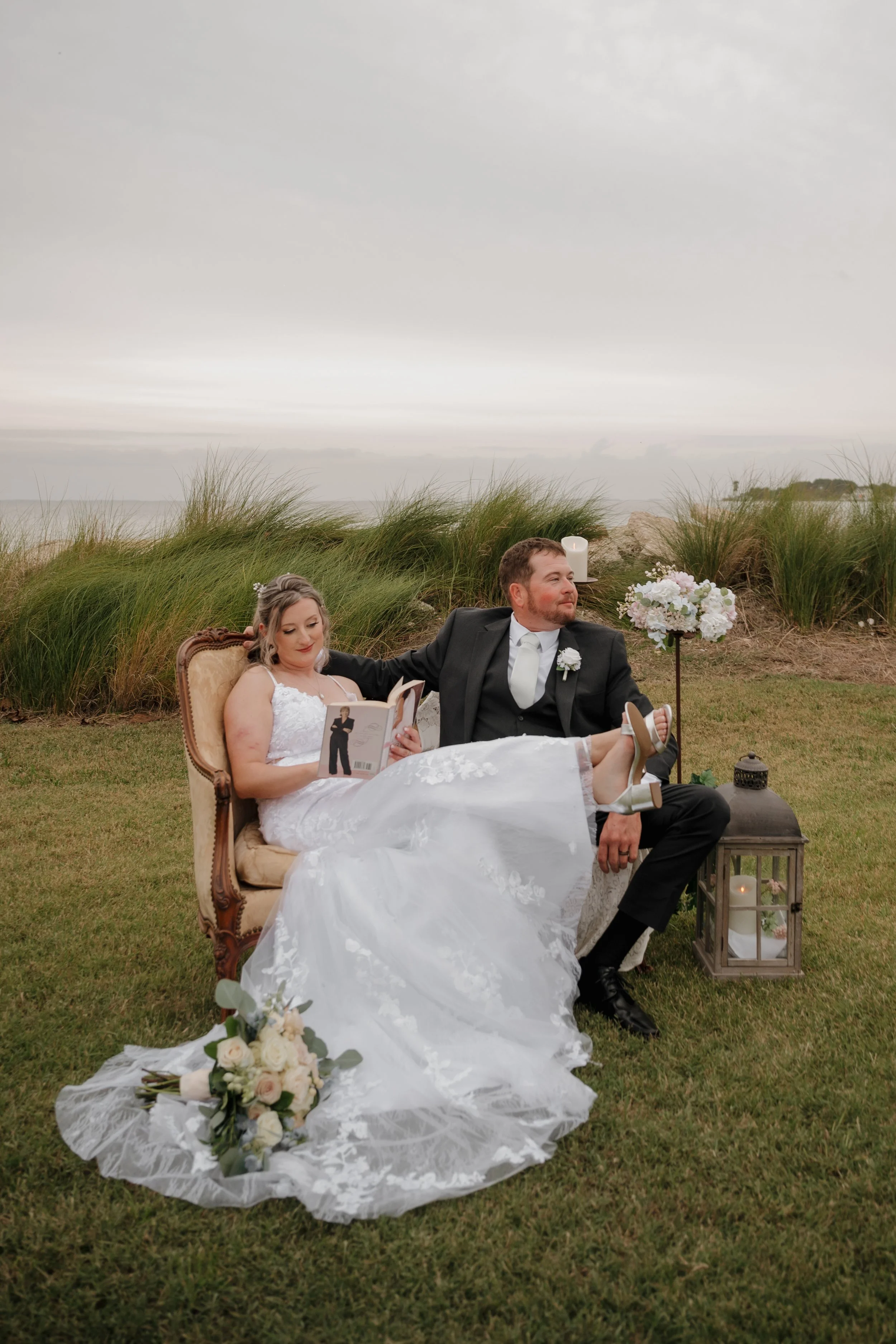 A newlywed couple sitting on a vintage sofa outdoors on a grassy lawn with a scenic ocean view and cloudy sky in the background. The bride is reading a book while the groom has his arm around her, and they are dressed in wedding attire with floral de