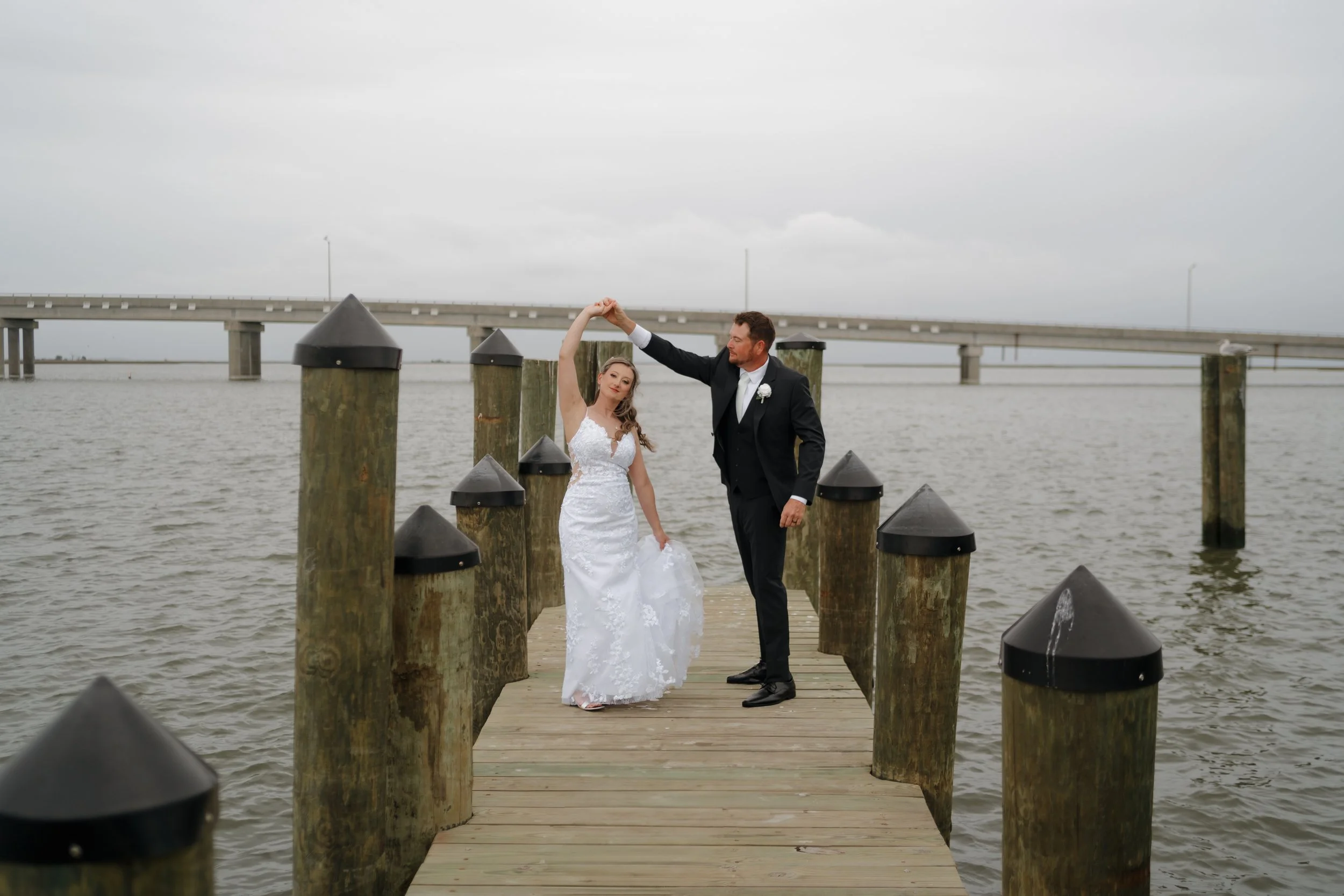 A bride and groom dance on a wooden dock over water with a bridge in the background, the groom twirling the bride.