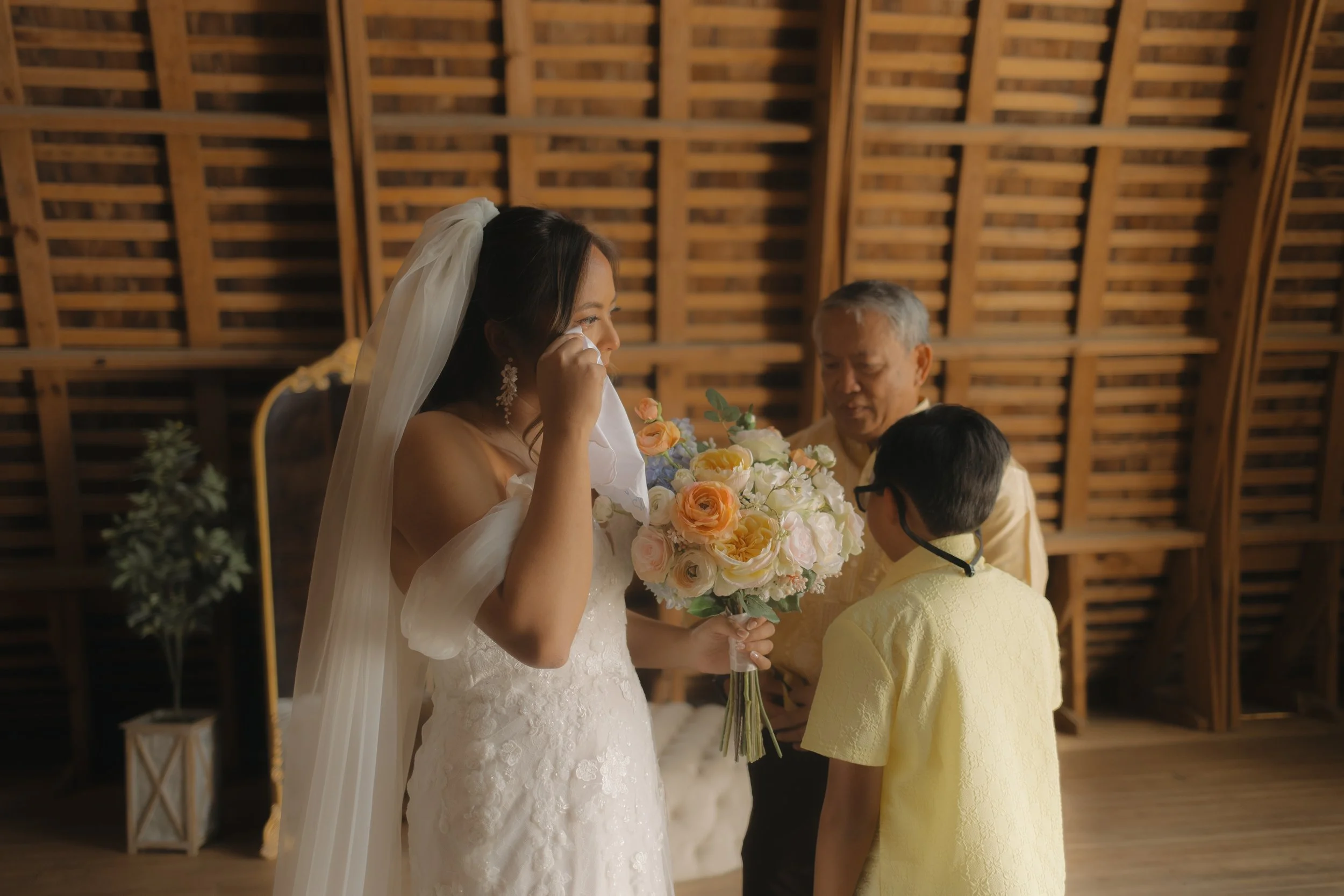 A bride in a white wedding dress with a veil is crying and holding a bouquet of flowers, while a young boy in a yellow shirt with glasses and two older men are standing next to her in an indoor setting with wooden wall panels.