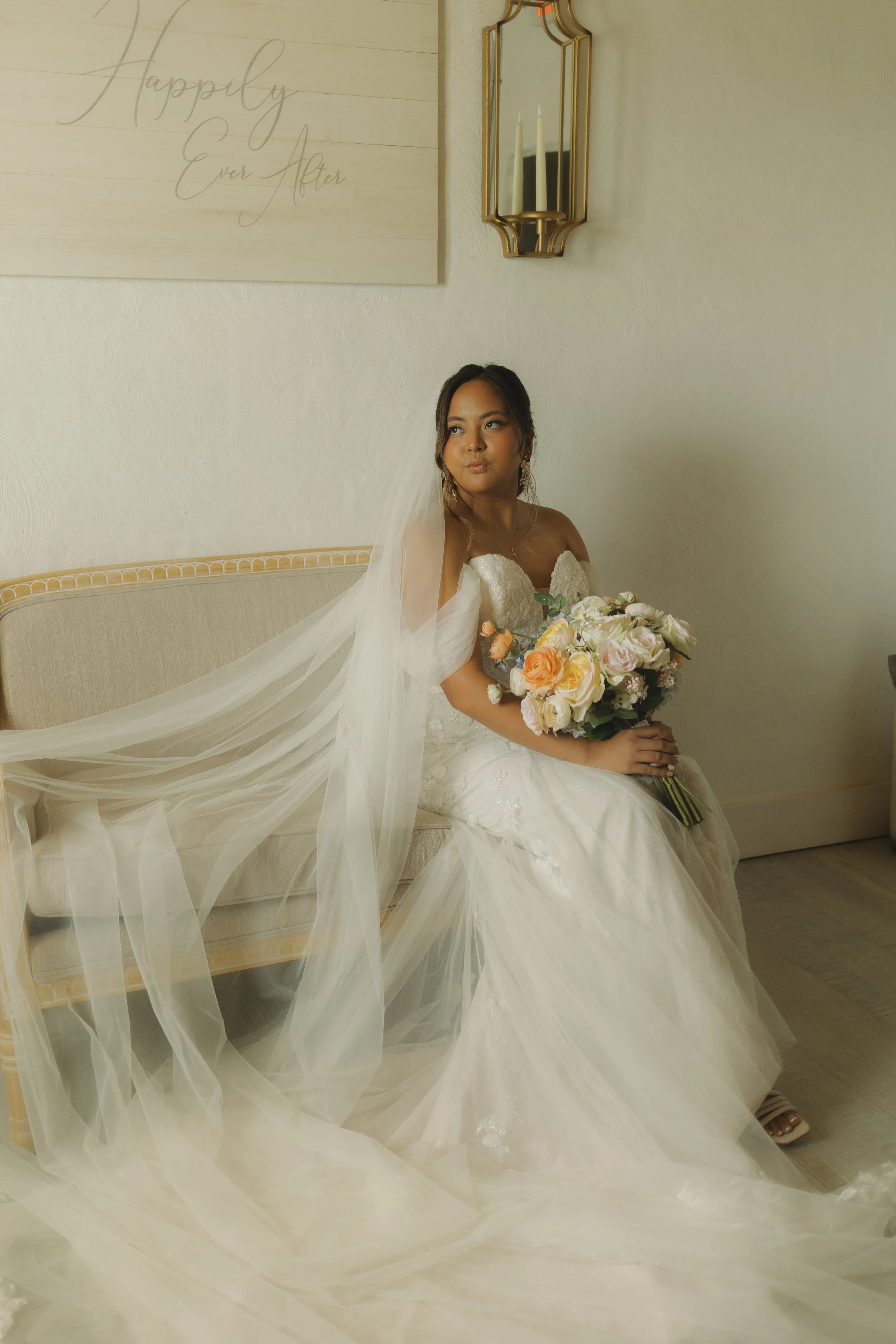 A bride in a white wedding gown sitting on a beige vintage sofa, holding a bouquet of pink, peach, and white flowers, with wedding veil cascading over her shoulders and onto the floor, in a room with a cream wall, gold-framed mirror, and a sign that 