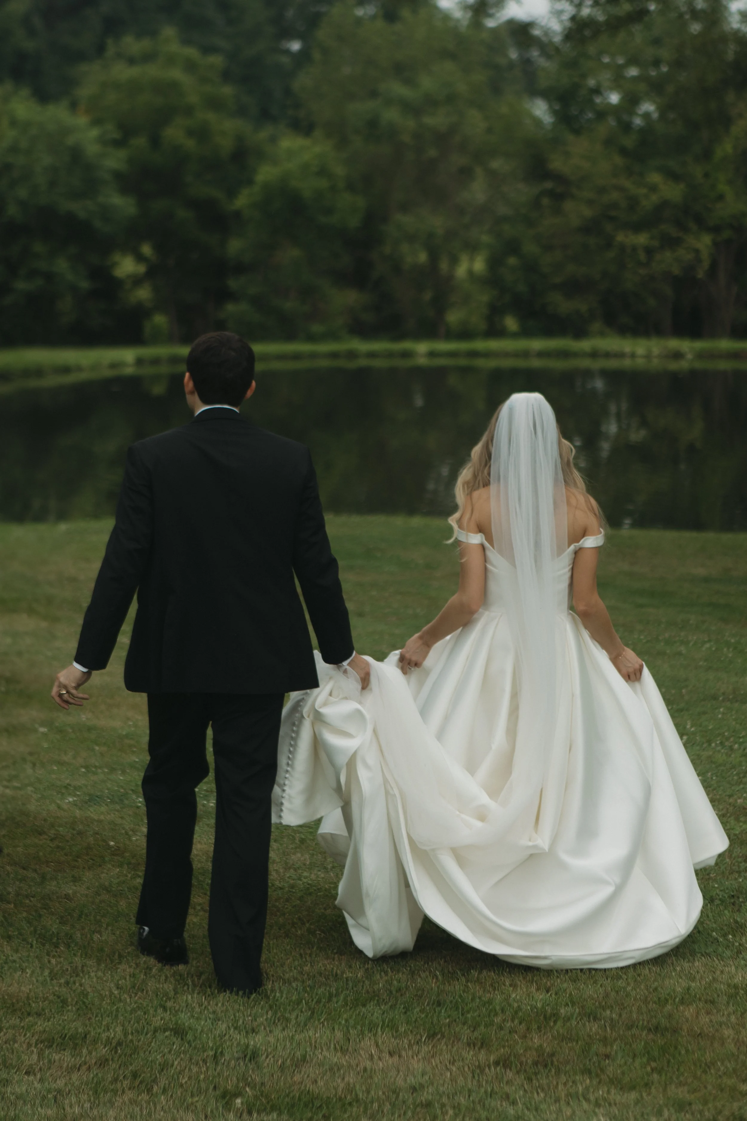 A bride and groom walking hand in hand towards a lake surrounded by green trees, with the bride wearing a white wedding gown and a veil, and the groom in a black suit.