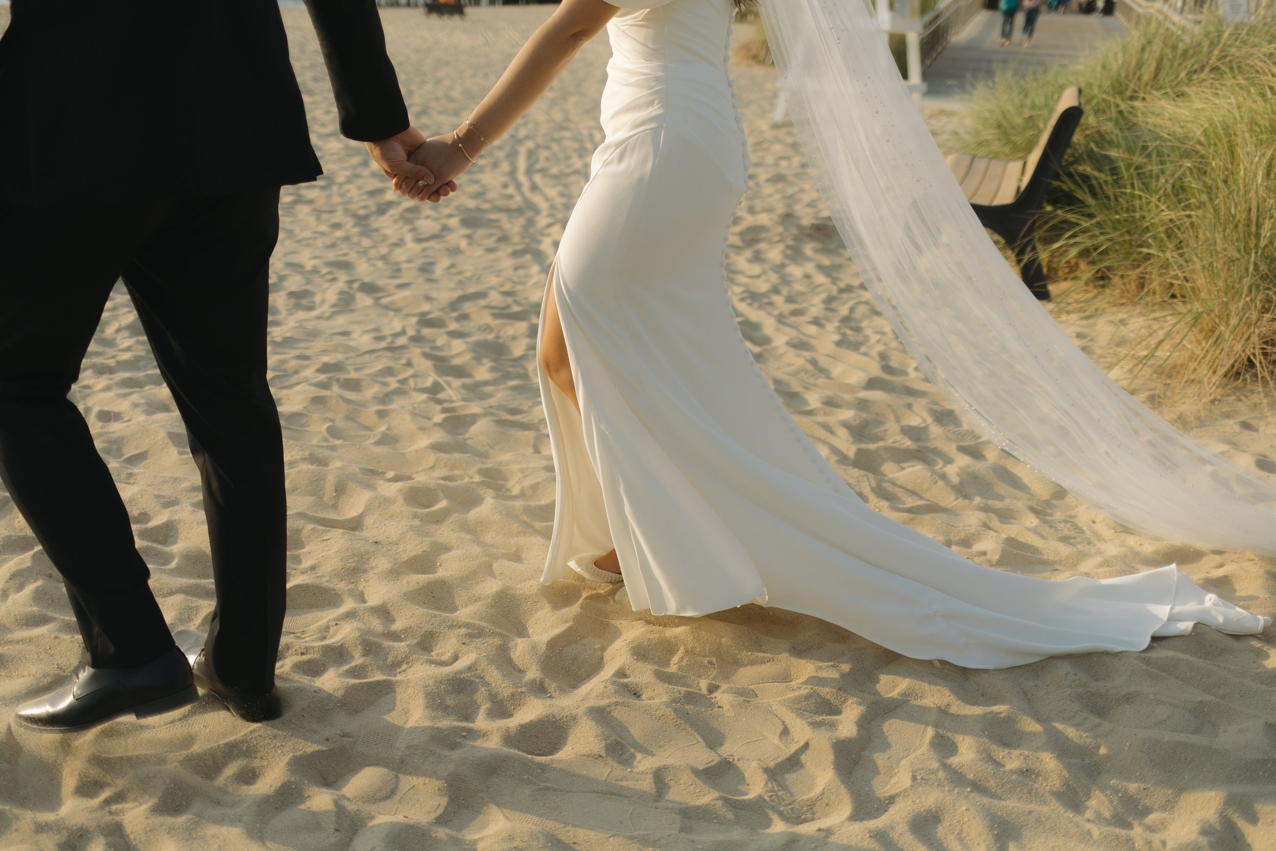 Couple holding hands on the beach, the woman wearing a white wedding dress and the man in a black suit.