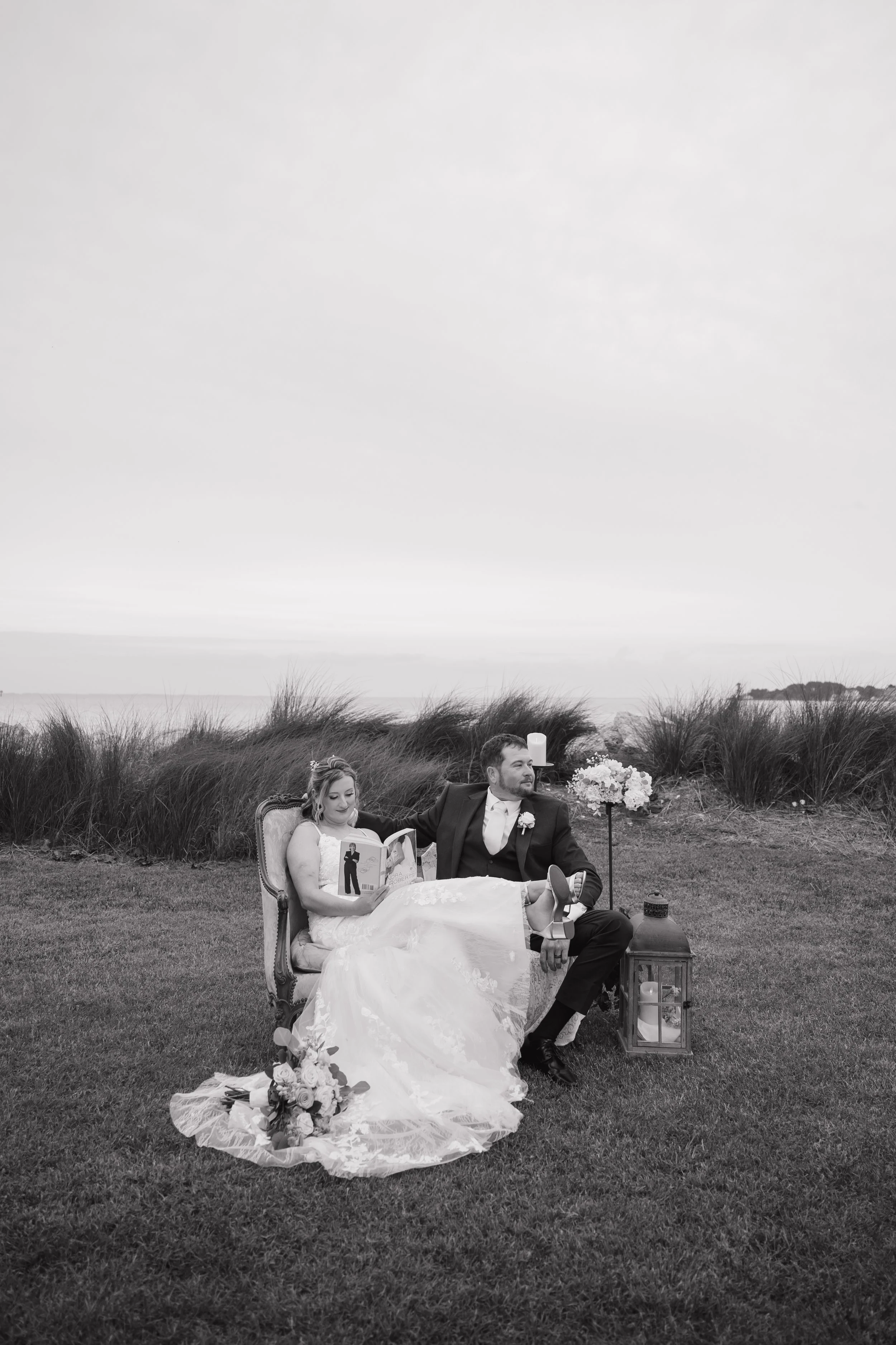 Black and white photo of a bride and groom sitting on a vintage sofa outdoors near the water, with the bride reading a wedding magazine, surrounded by flowers, candles, and lanterns.