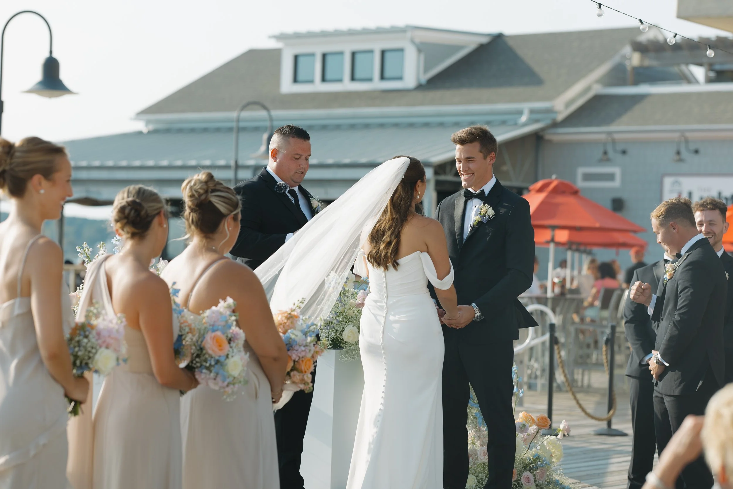A wedding ceremony taking place outdoors on a sunny day, with a bride and groom holding hands, surrounded by bridesmaids and groomsmen.
