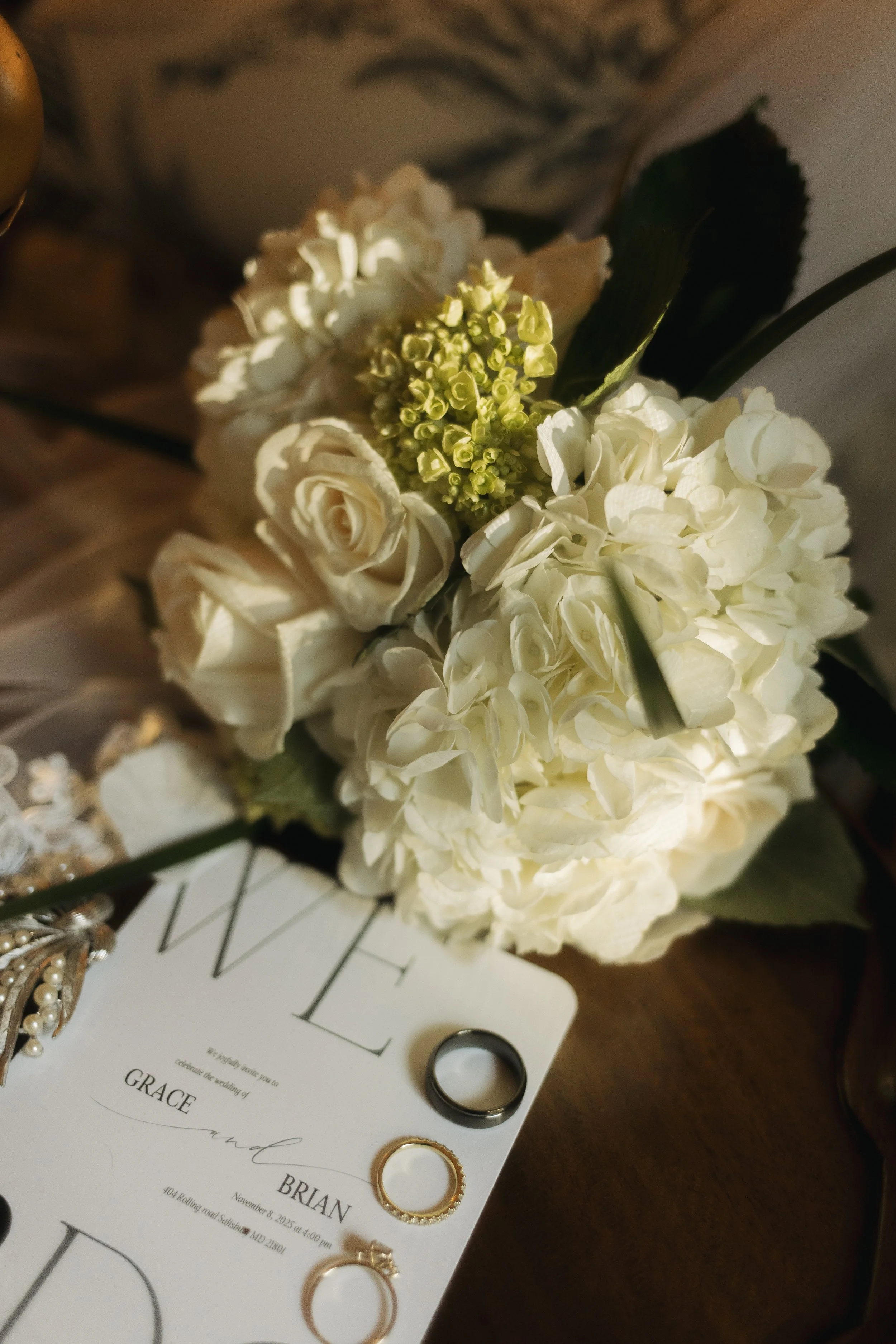 A bouquet of white flowers including roses and hydrangeas, with wedding rings placed on a wedding invitation card that has the names Grace and Brian, on a wooden surface.
