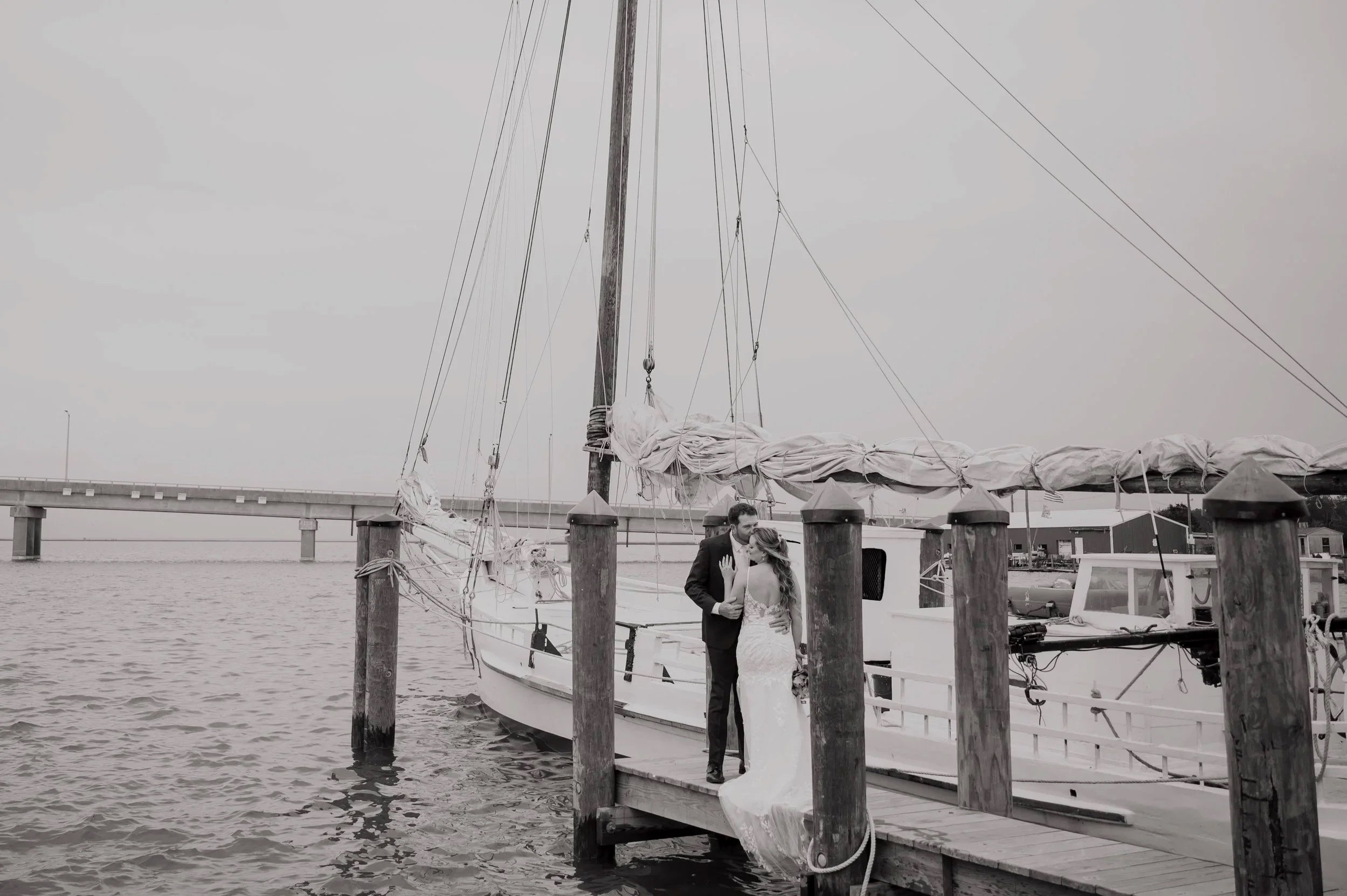 A bride and groom sharing a kiss on a dock with a sailboat. The bride is wearing a white wedding dress, and the groom is dressed in a black suit. Sailboat masts and a bridge are visible in the background.