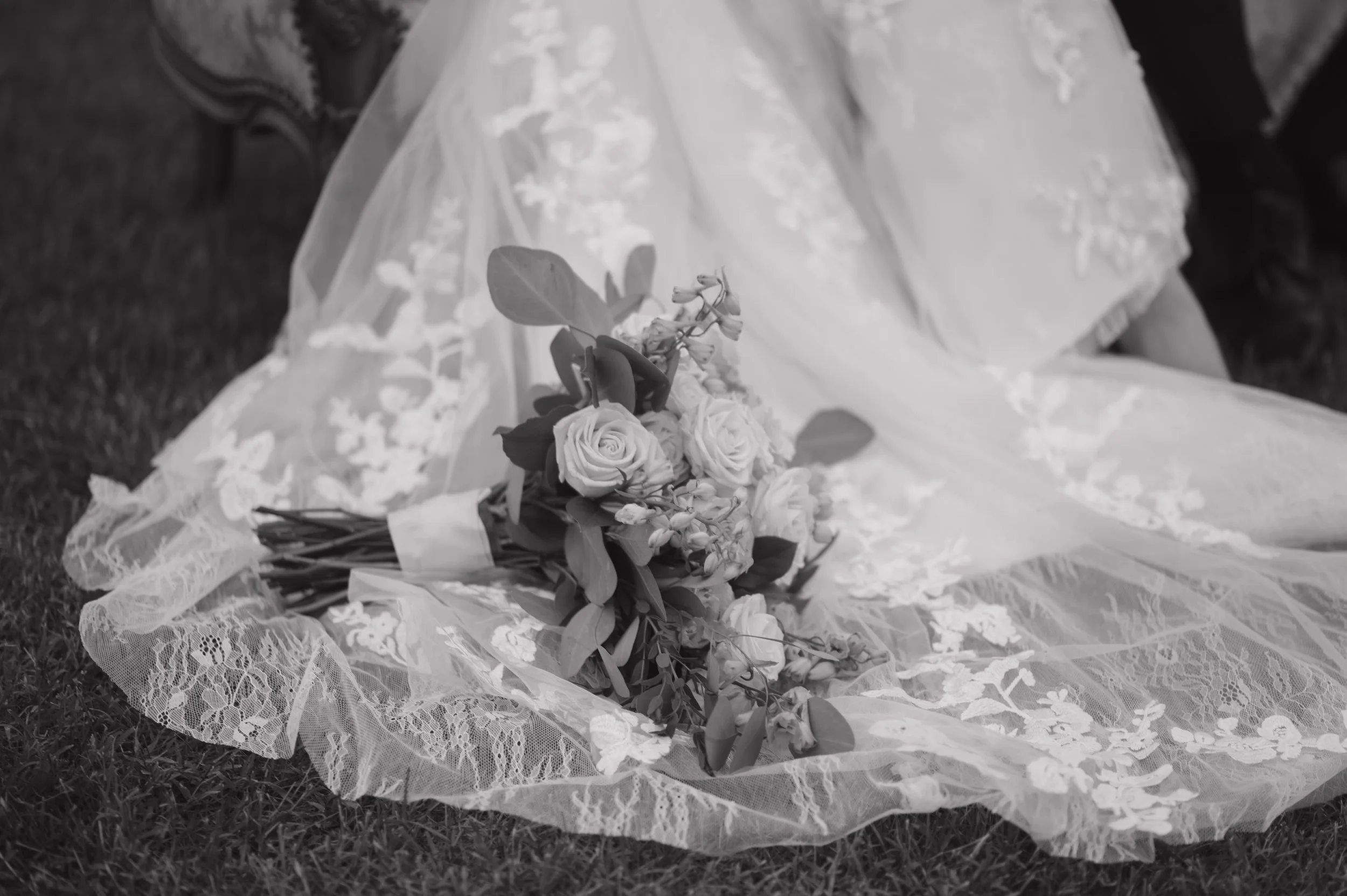 A bouquet of roses and greenery lies on a lace fabric train of a wedding dress, with a person seated on the grass nearby.