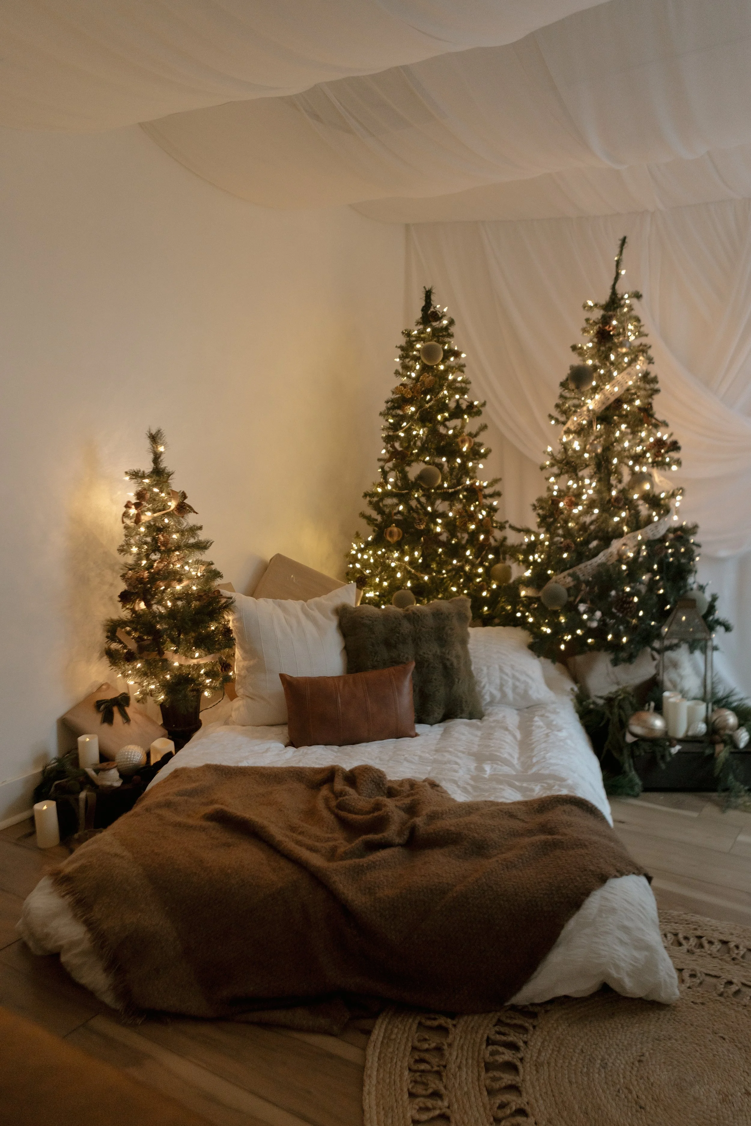 A cozy bedroom with Christmas trees decorated with lights and ornaments, a bed with pillows and a brown blanket, candles, and holiday decorations.