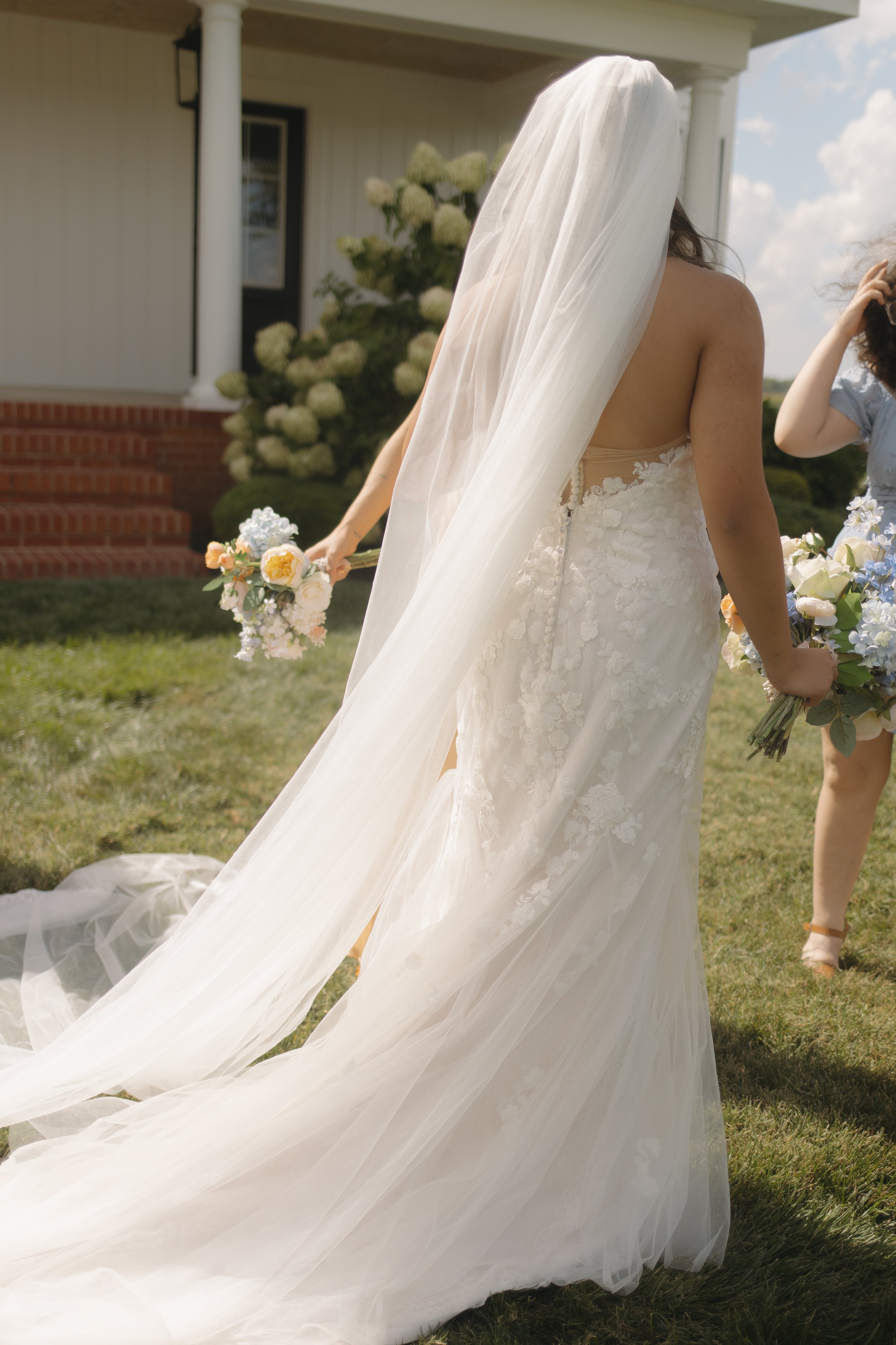 Bride in a white wedding dress with a long veil, holding a bouquet of flowers, standing on a grassy lawn outside a house.