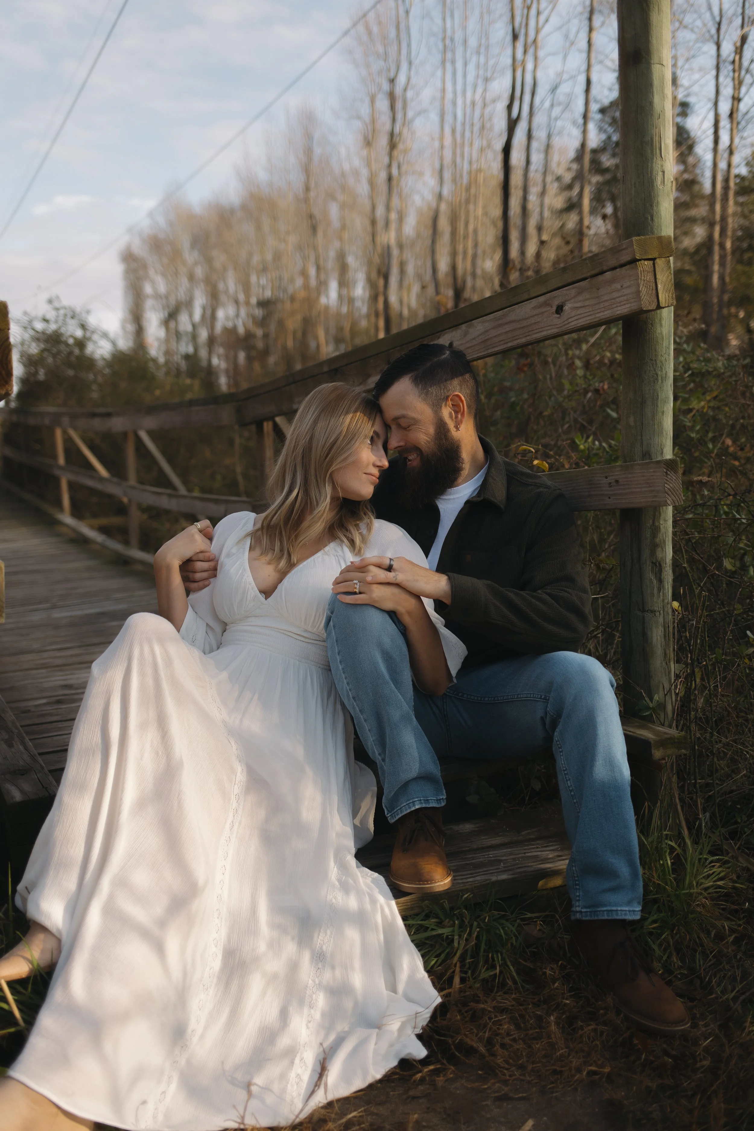 A couple sits closely together on a wooden bridge in a wooded area, sharing a tender moment. The woman wears a white dress and is leaning into the man, who has a beard, dark hair, and wears a black jacket and blue jeans. They are touching foreheads and smiling softly.