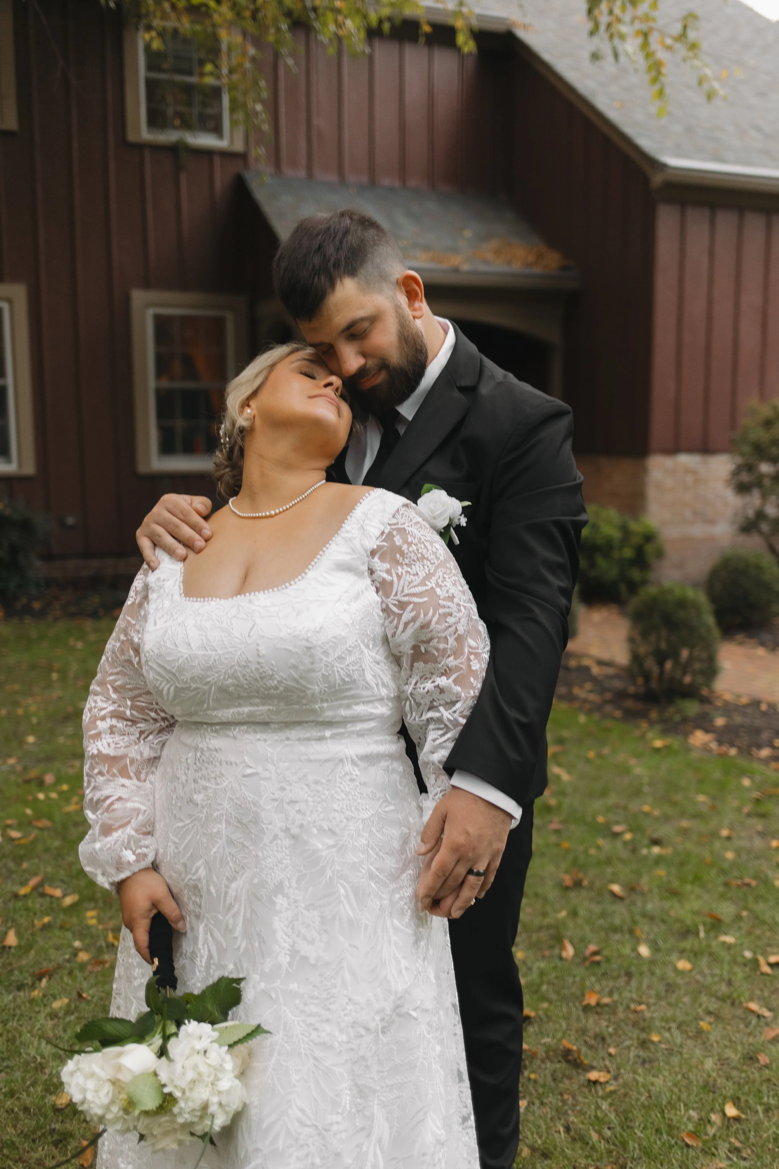 A bride in a white lace wedding dress and pearl necklace leans into a groom in a black suit with a white shirt and boutonniere, as they embrace outdoors near a brown house with trees and bushes.
