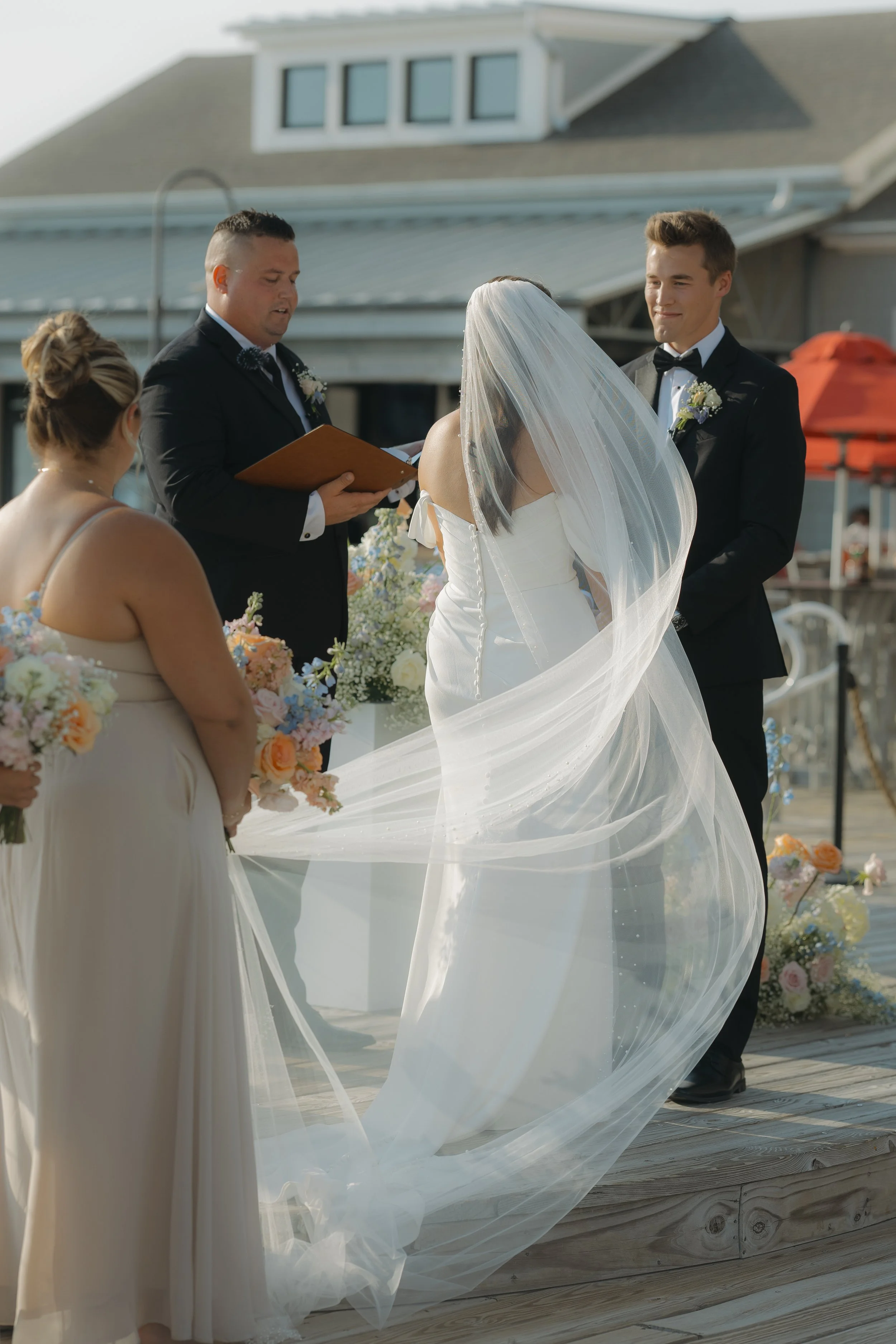 A wedding ceremony taking place outdoors on a wooden deck, with a bride in a white dress and veil, an officiant, and two bridesmaids holding bouquets of flowers.