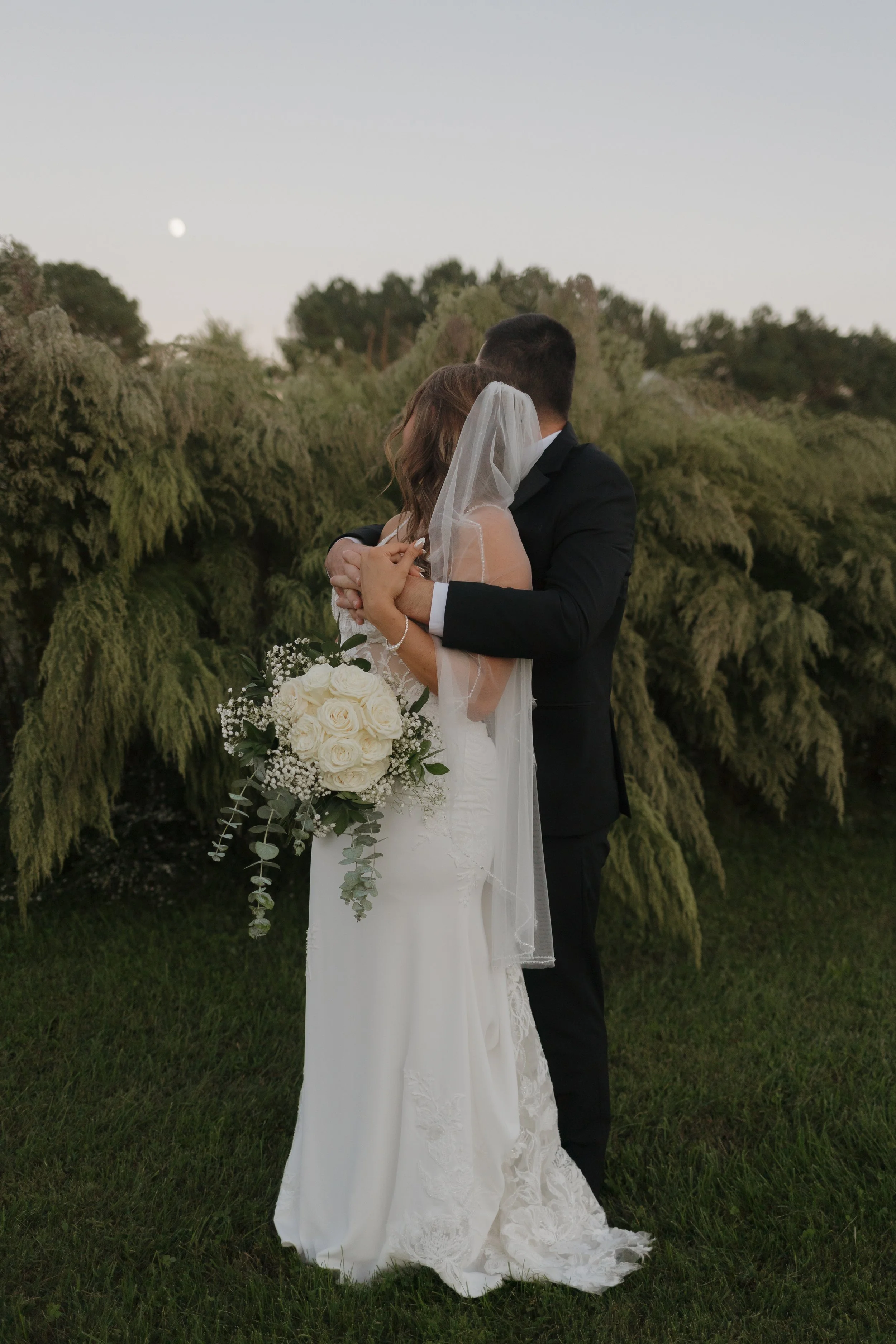 A bride and groom hugging outdoors at sunset, with the moon visible in the sky, in front of lush green trees.