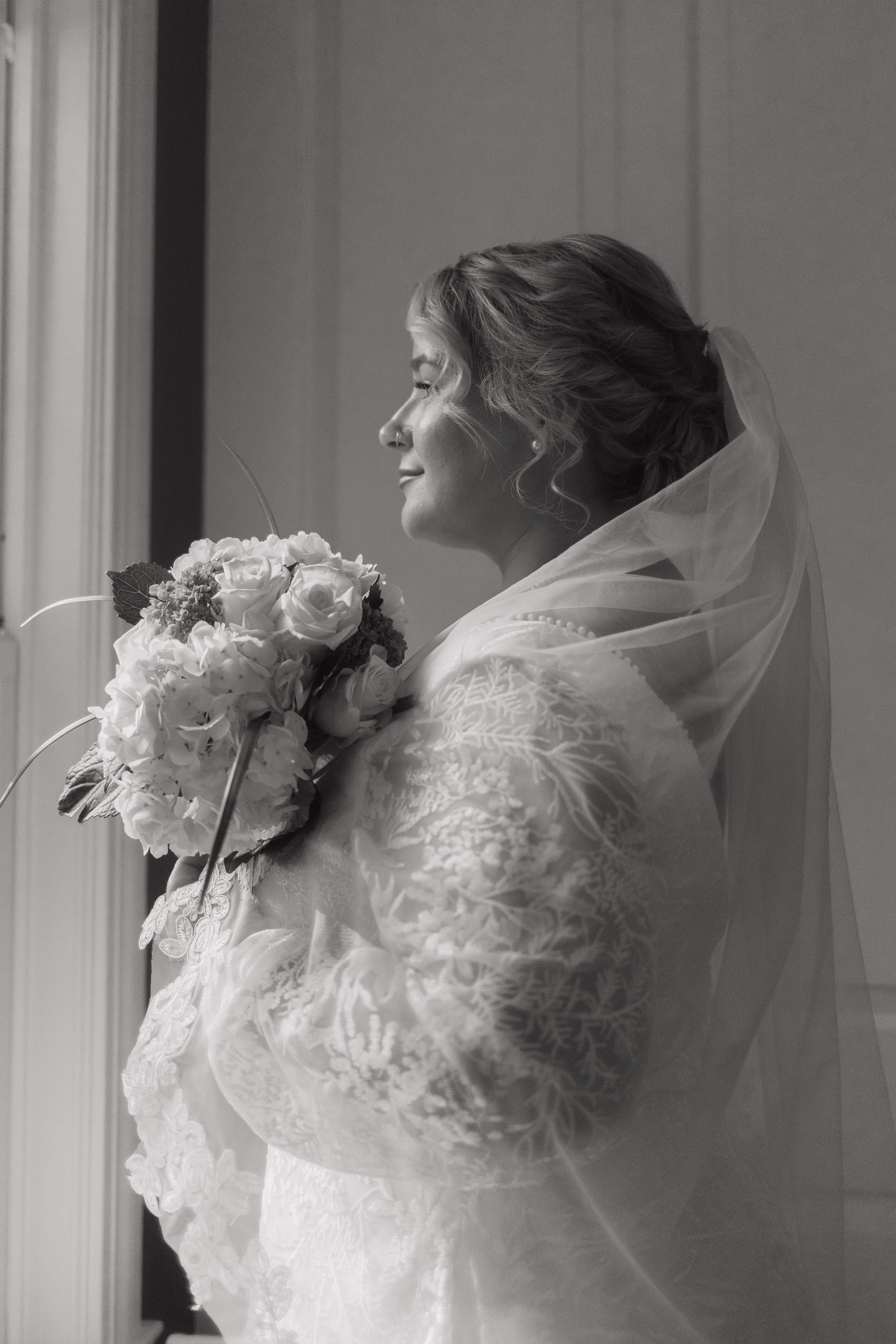 Black and white photo of a bride holding a bouquet, wearing a lace dress and veil, with her eyes closed and a peaceful smile.
