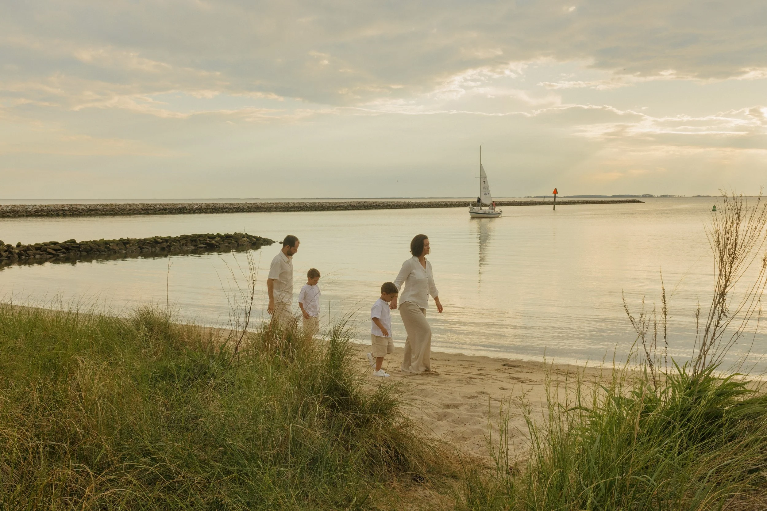A family of four, dressed in white and beige, walking on a sandy beach near the water at sunset. A sailboat is sailing in the distance, and a breakwater extends into the water. Sea grasses are in the foreground.