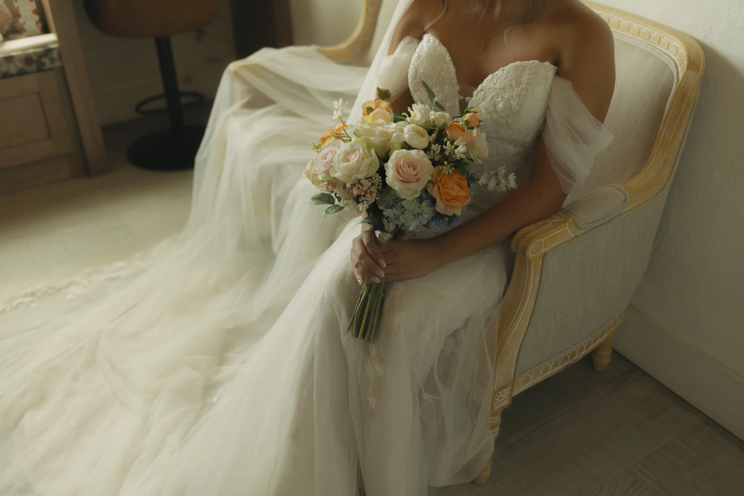 A bride in a white wedding dress sitting on a beige armchair, holding a bouquet of flowers with roses in light pink, peach, and white, and other mixed flowers.