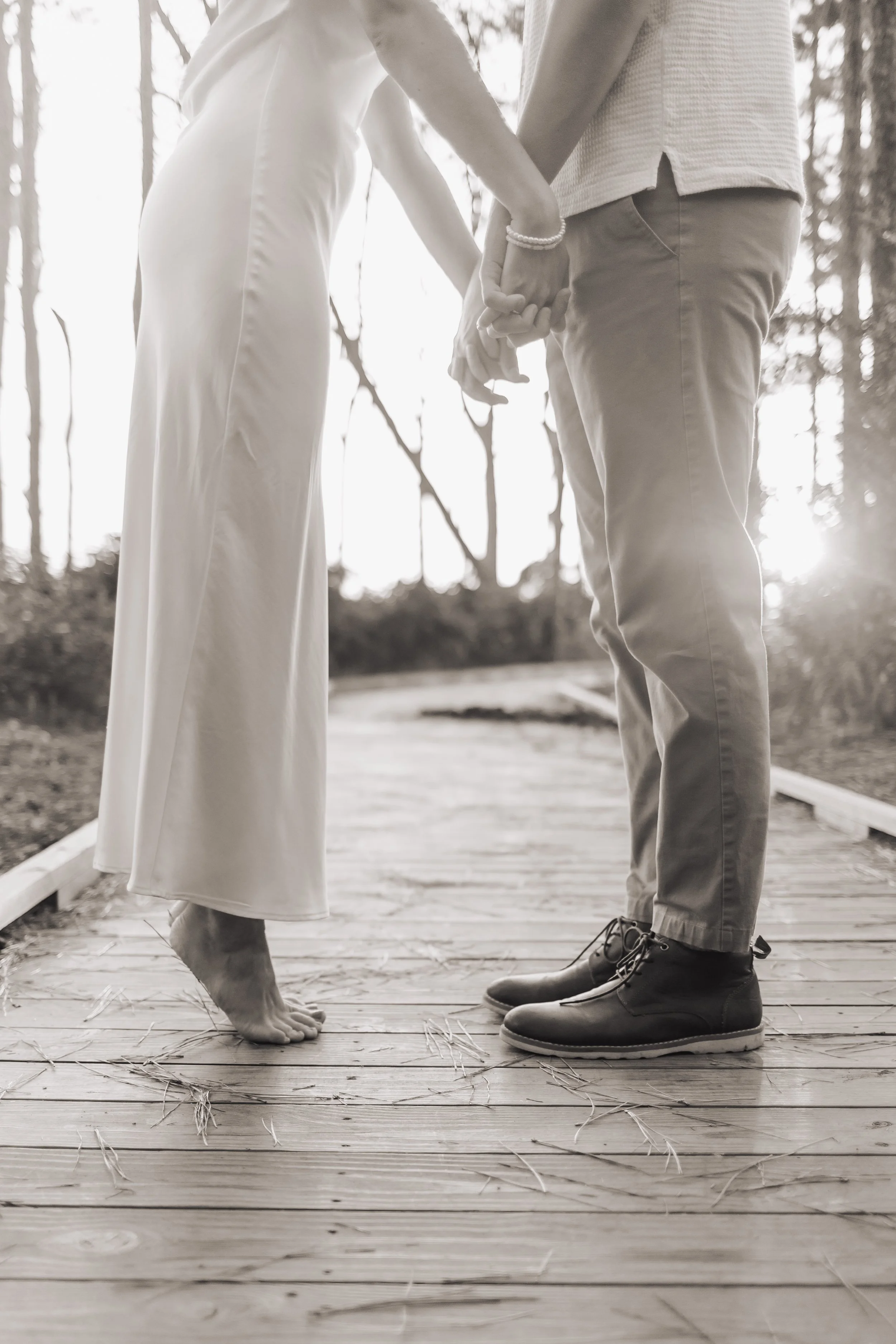 A couple holding hands on a wooden bridge, with the woman's feet visible, barefoot, and the man's wearing black boots, during sunset.