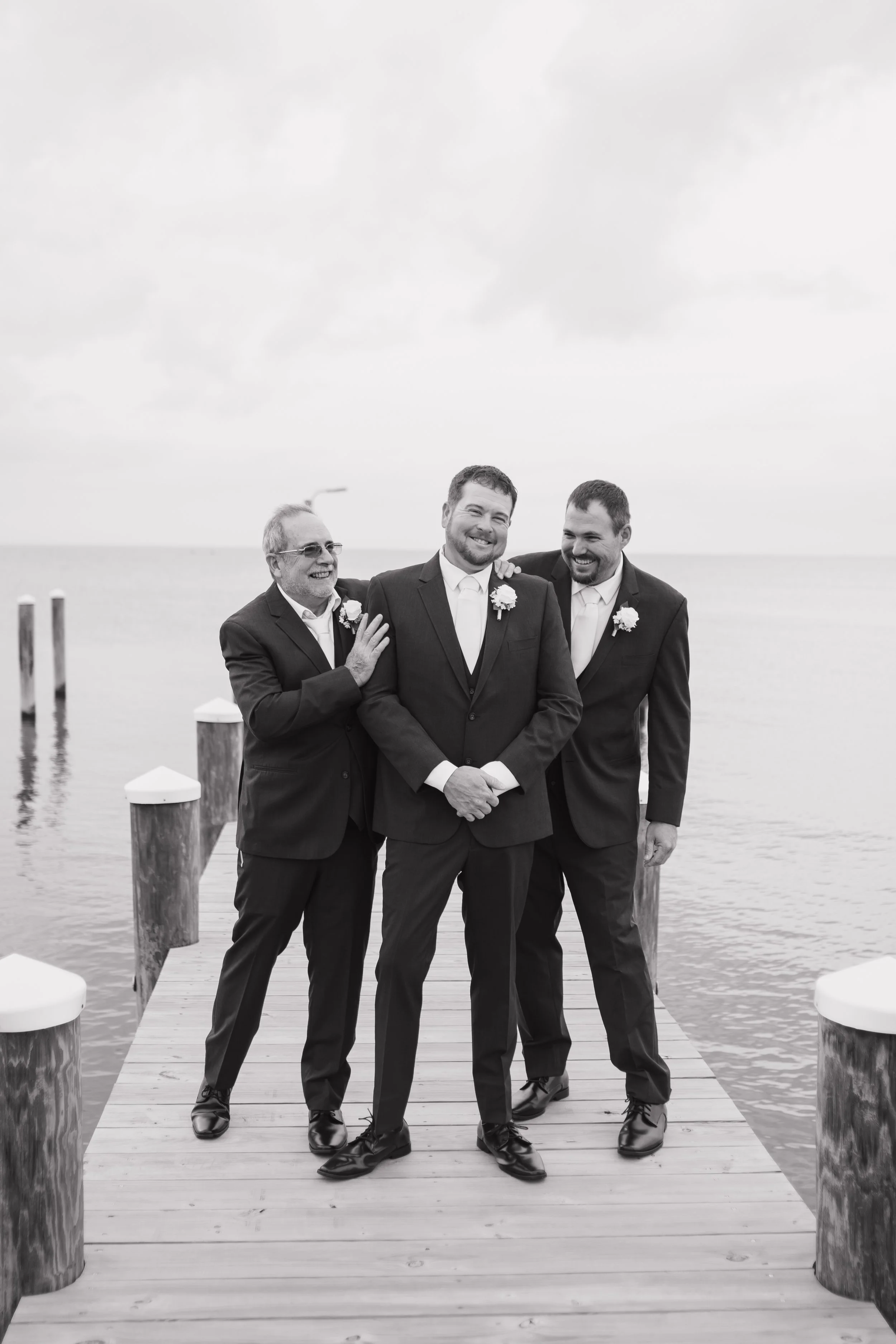 Three men in suits standing on a wooden dock by the water, smiling, with one man having his arm around another, in a black and white photo.
