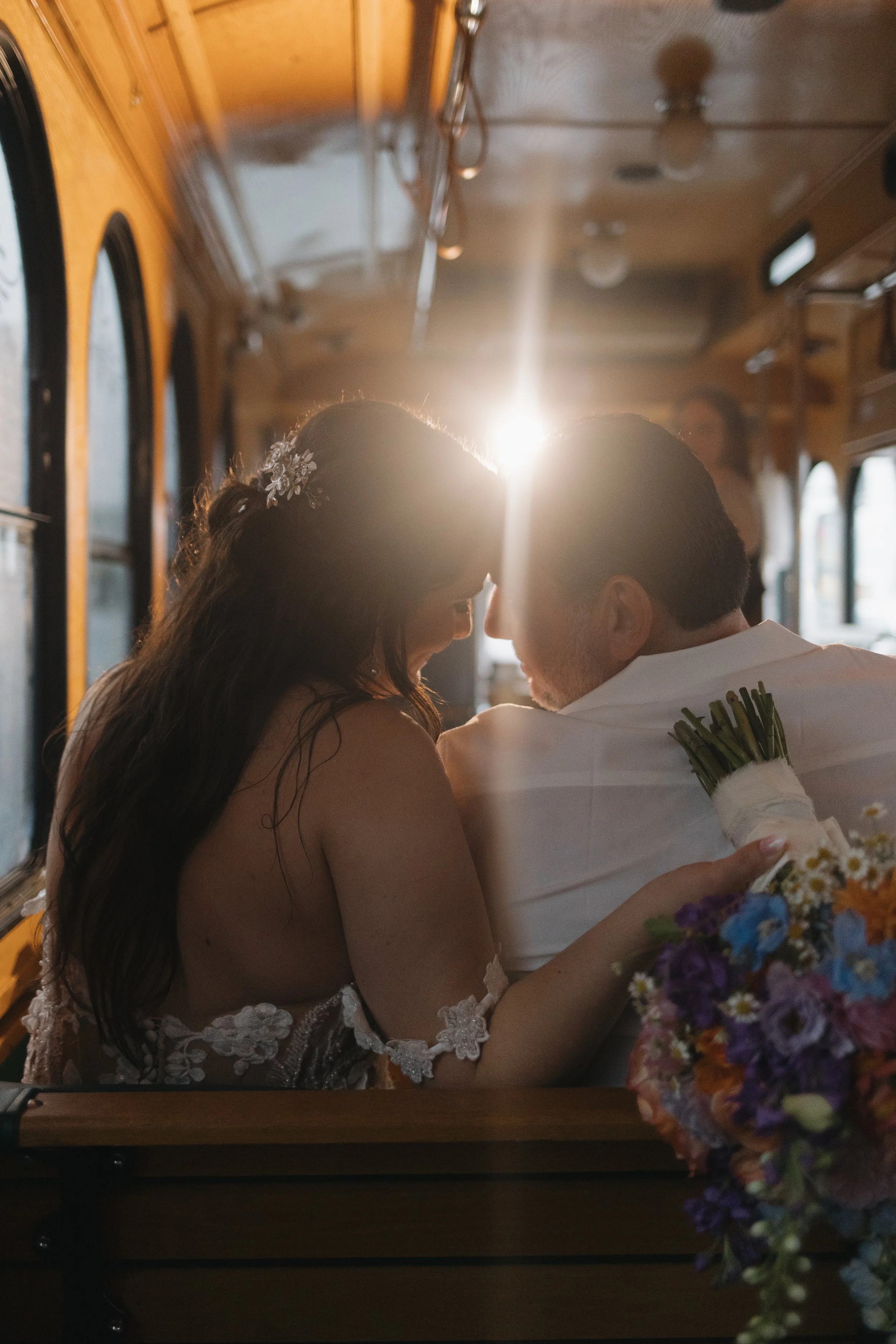 A bride and groom sitting closely together on a trolley, sharing a tender moment with their foreheads touching and smiling. The bride has long, dark hair with a floral accessory. The groom has short hair and a trimmed beard. They are holding a bouque