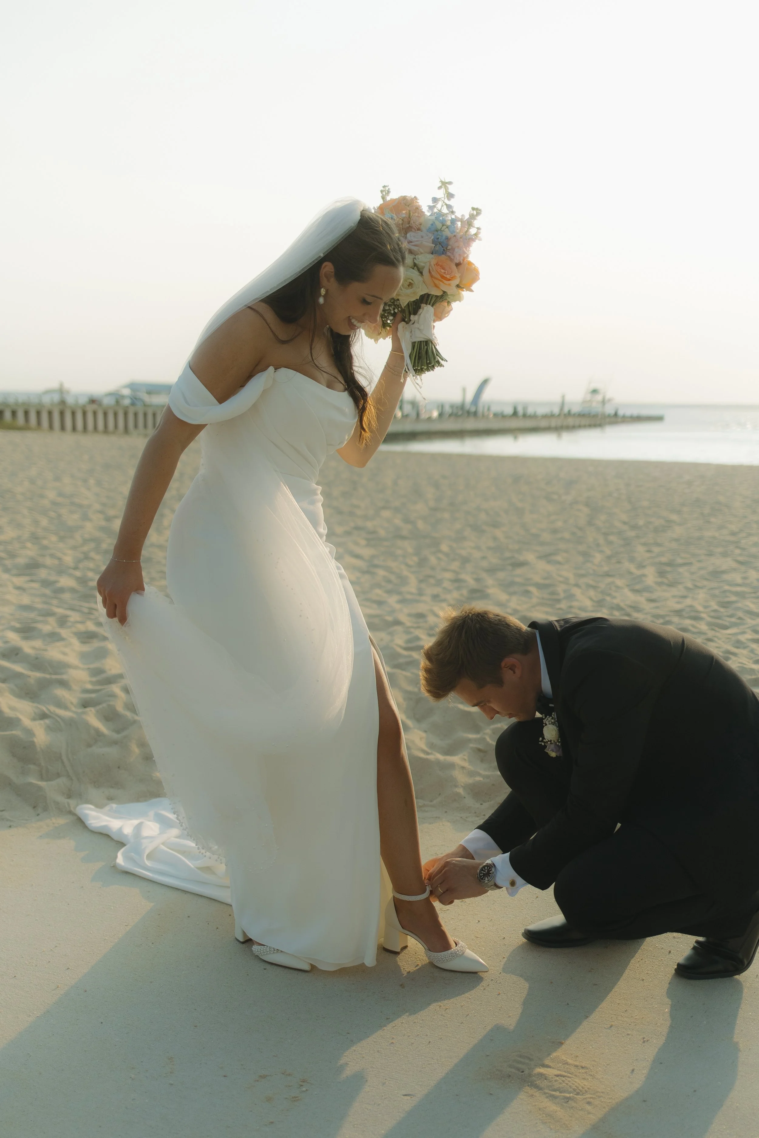 A bride in a white wedding dress adjusting her shoe while a groom in a black suit helps her on the beach during sunset.