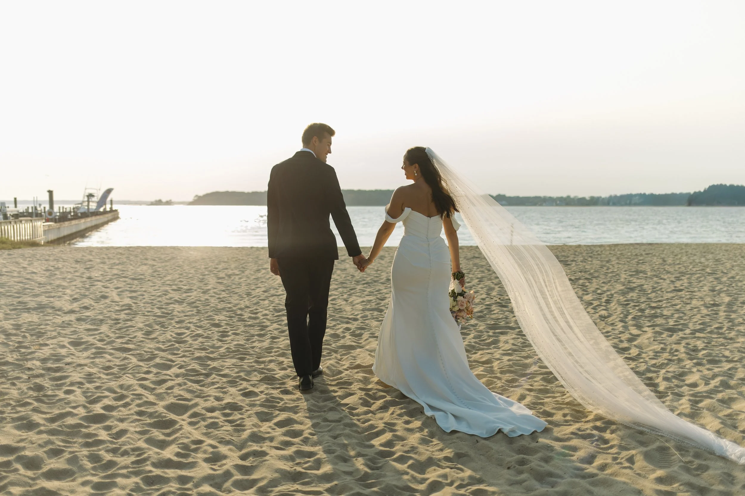 A bride and groom holding hands on a sandy beach at sunset, with water and distant land in the background, celebrating their wedding.